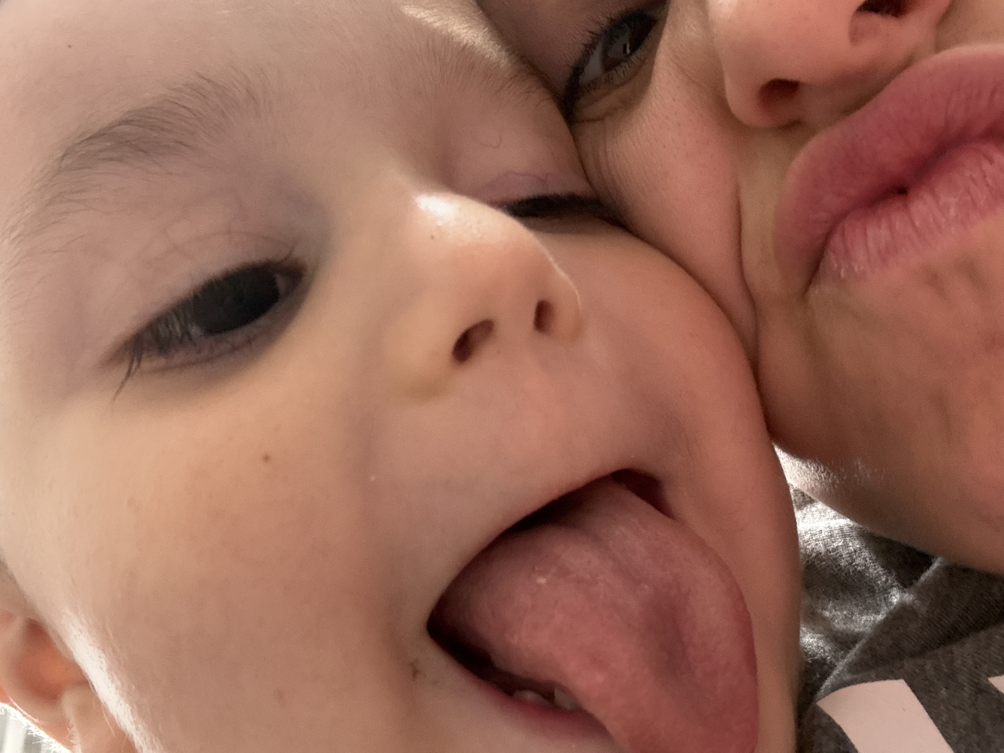 This is a close-up, low-angle, indoor photograph of two young children. The child in the foreground has their mouth open, sticking out their tongue. The child behind them has their eye visible, looking toward the camera. The image is tightly framed, focusing on the faces of the children. The background is dark and out of focus. The location of this image is the City of Yonkers, United States of America.