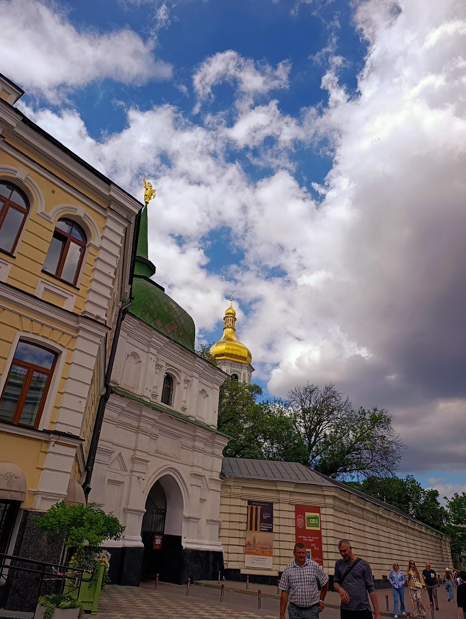 The image shows a view of St. Sophia's Cathedral in Kyiv, Ukraine, on a partly cloudy day. In the foreground, a section of a yellow building with white architectural detailing and arched windows is visible on the left, angled towards the viewer. A green-domed structure with a golden angel statue on its spire is behind and to the right of the yellow building. Further in the background, a golden dome of another church structure is visible. People are walking on a paved walkway in front of a long, beige, stone wall with posters on it. The sky above is a mix of bright blue with scattered white clouds and darker, more ominous clouds.