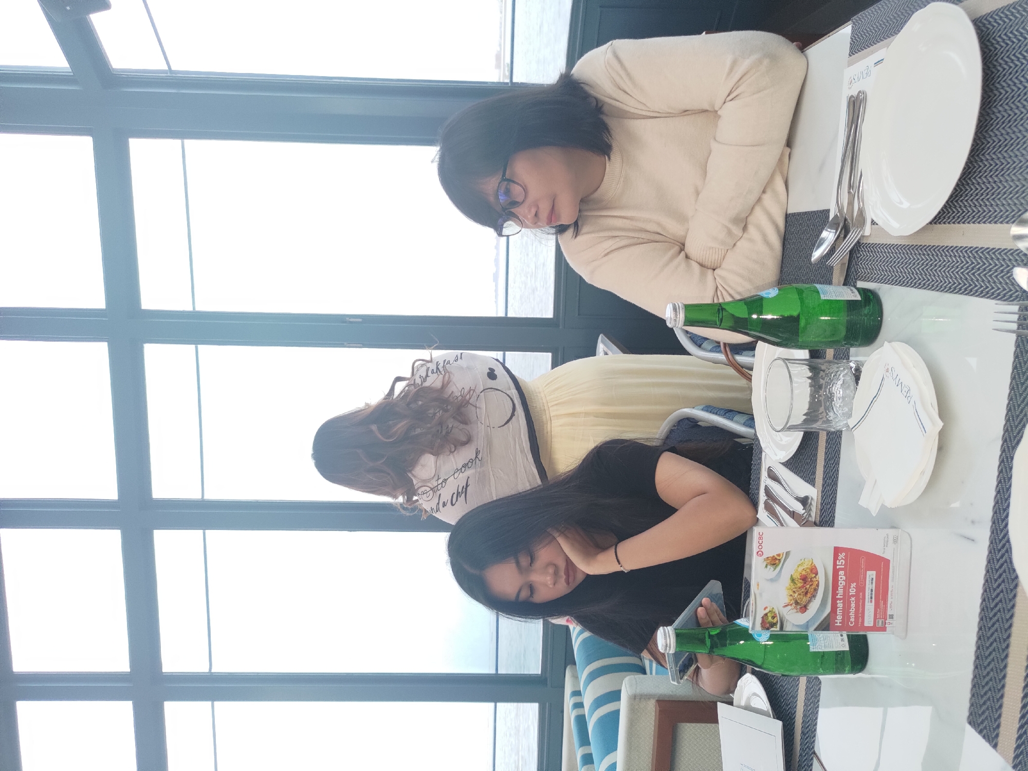 Three women are seated at a table in a restaurant with a view of the sea through large windows. The woman in the foreground on the left is looking down at her phone, resting her chin on her hand. She has long, dark hair and is wearing a black t-shirt. To her right, a green bottle of water and a promotional flyer are on the table. The woman in the foreground on the right is wearing glasses and a beige sweater, looking to the side. Her arms are crossed. Behind them, a third woman, facing away from the camera, is wearing a long yellow skirt and a scarf draped over her shoulders. The table is set with plates, cutlery, and glasses. The environment appears to be a modern restaurant.