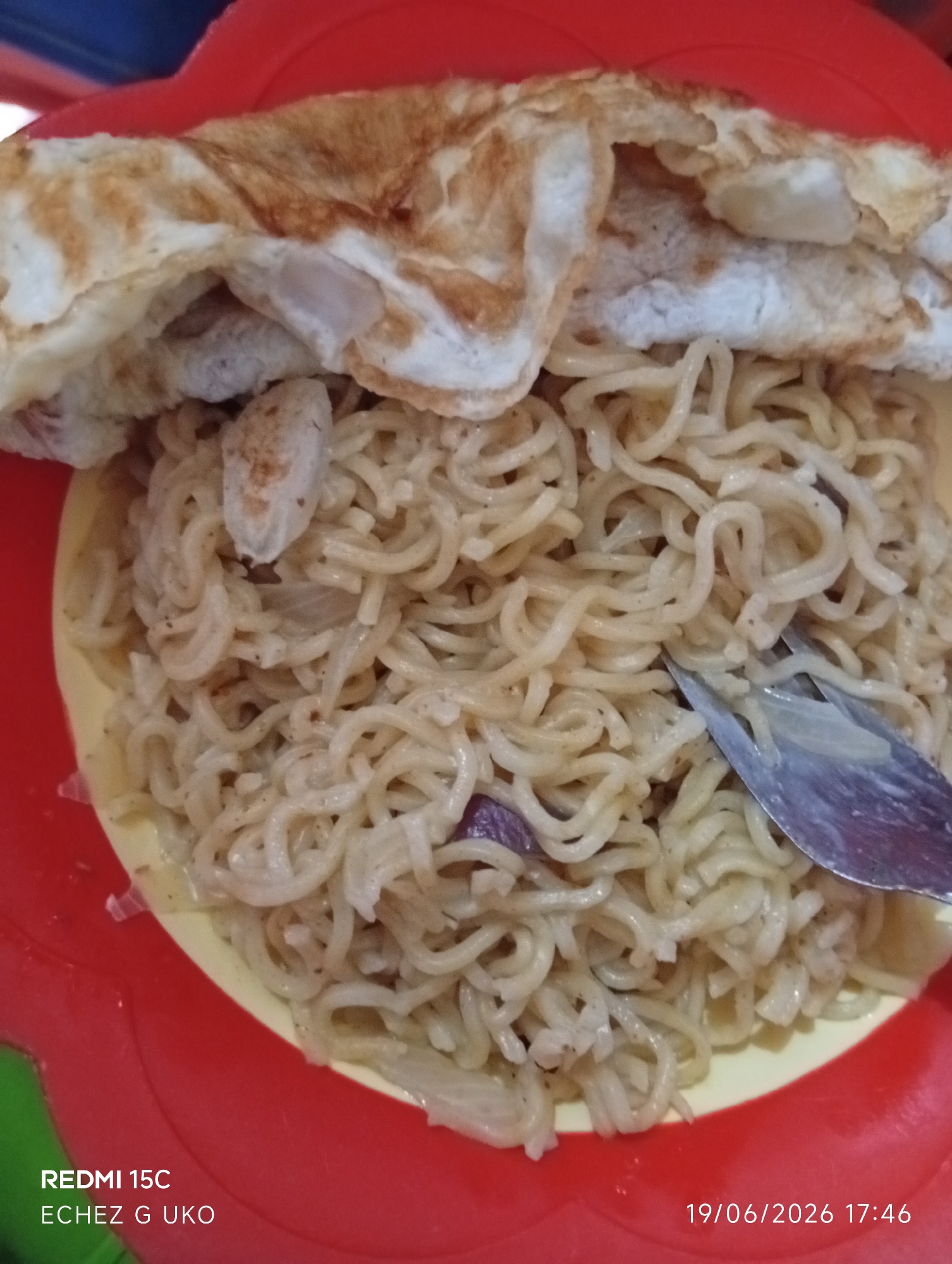 In the city of Jalingo, Nigeria, a close-up shot captures a red plastic plate filled with noodles and topped with a folded omelet. A fork is visible, partially submerged in the noodles, suggesting the meal is being eaten. The noodles appear to be a pale yellow or off-white color, with some pieces of chopped onion mixed in. The omelet is golden brown with some lighter, fluffy sections. The background is out of focus, but hints of blue and green can be seen, possibly indicating a table and an outdoor setting. The timestamp "19/06/2026 17:46" is visible in the lower right corner, along with "REDMI 15C" and "ECHÉZ G UKO" in the lower left.