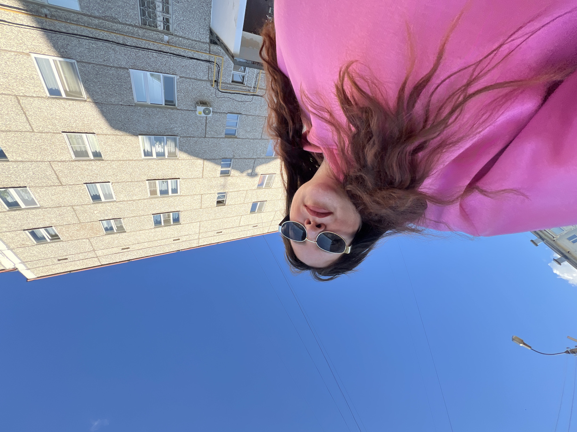 A woman with long brown hair, wearing sunglasses and a bright pink hoodie, is pictured outdoors in Noviy Urengoy, Russia. The photograph is taken from a low angle, with the woman positioned in the upper portion of the frame. Below her is a clear blue sky. In the background, a gray concrete apartment building is visible. The setting appears to be a sunny day.