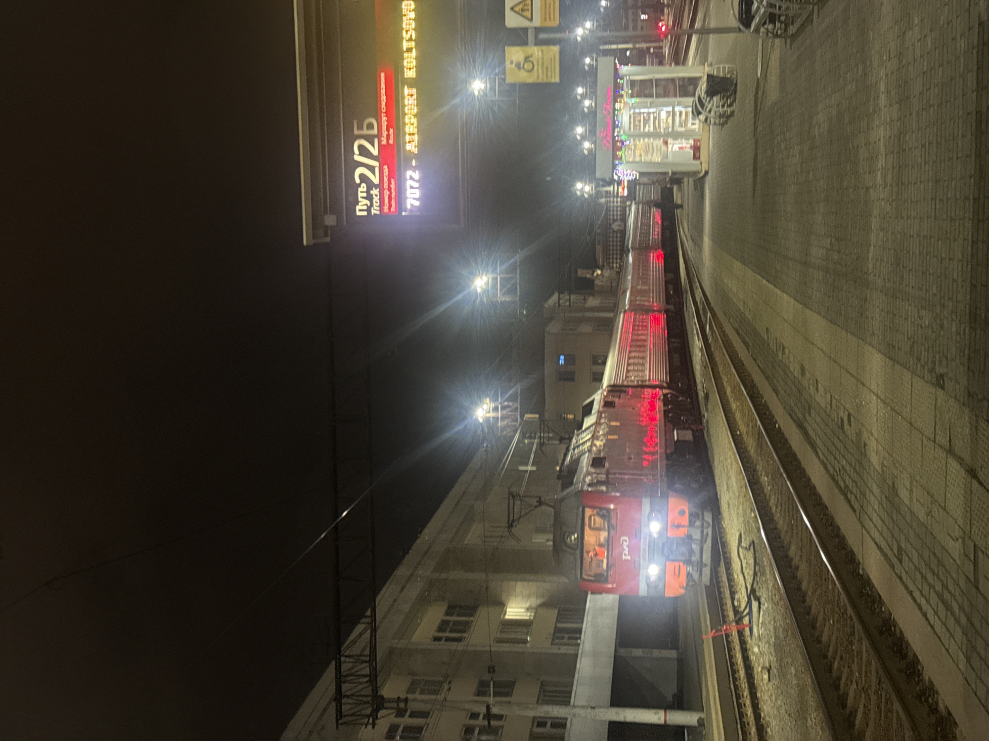 The image shows a train stopped at a train station platform in Yekaterinburg, Russia, at night. The train is red and has headlights illuminated. The platform is made of grey concrete. A trackside sign reads "Путь 2/2Б Track" followed by "7072 - AIRPORT KOLTSOVO." The sign indicates the train's destination is Koltsovo Airport, which is located in Yekaterinburg. There are overhead lights and some lit buildings in the background.