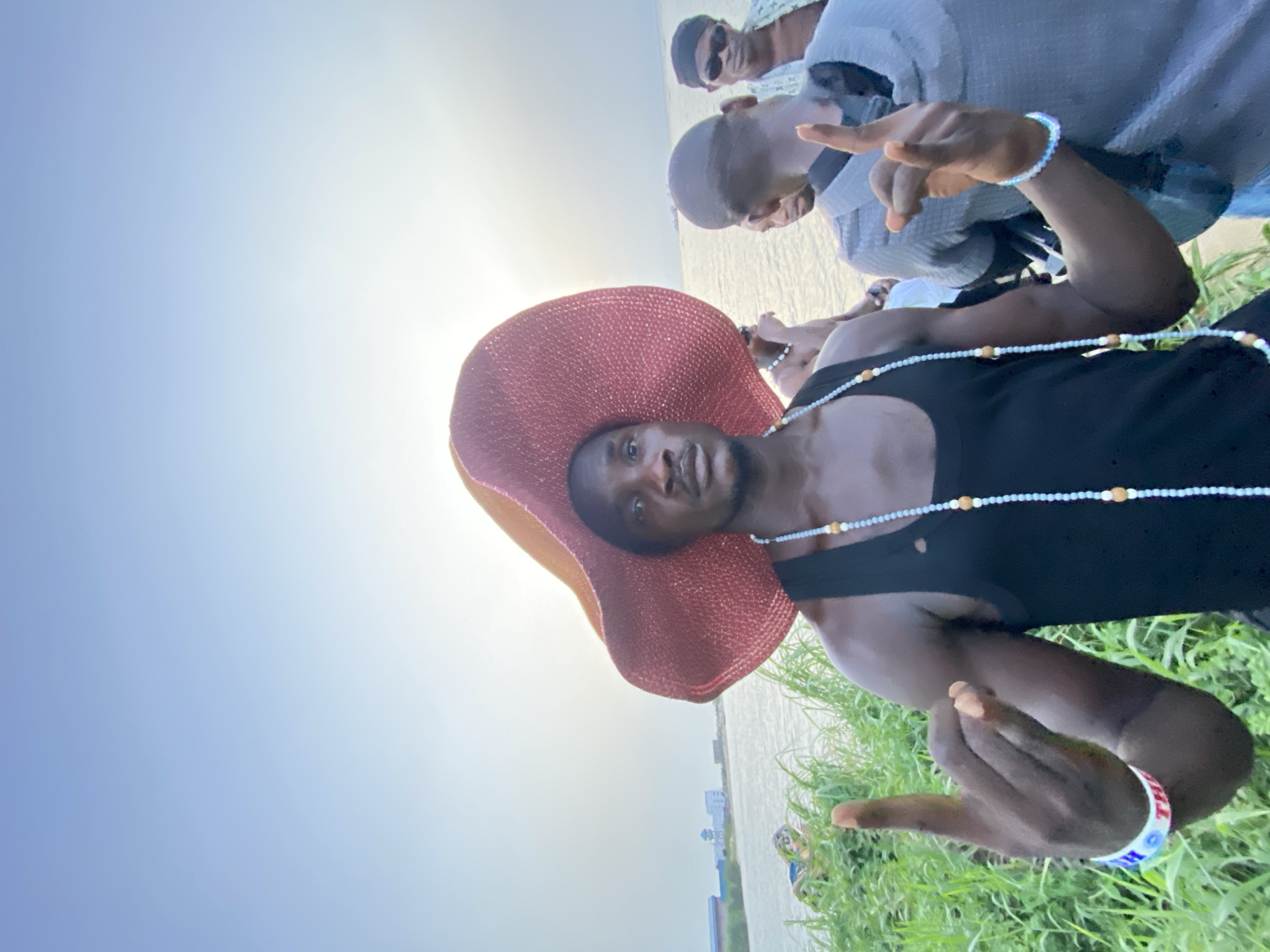 The scene is an outdoor setting near a body of water, likely a river or lagoon, with a backdrop of a pale sky and bright sunlight. Some buildings in the distance are suggestive of an urban or semi-urban environment. Green grass and vegetation are visible in the foreground.
In the foreground, a dark-skinned person is the main subject, wearing a black tank top, a beaded necklace, a bracelet with blue and white patterns, and a large, wide-brimmed reddish-brown hat. The person is gesturing with both hands.
Behind the main subject, there are at least two other individuals. One is wearing sunglasses and a patterned shirt. The lighting conditions suggest it is daytime. The specific location is not definitively confirmed as Ikorodu, Nigeria, based solely on the visual elements of the image, but Ikorodu is suggested.