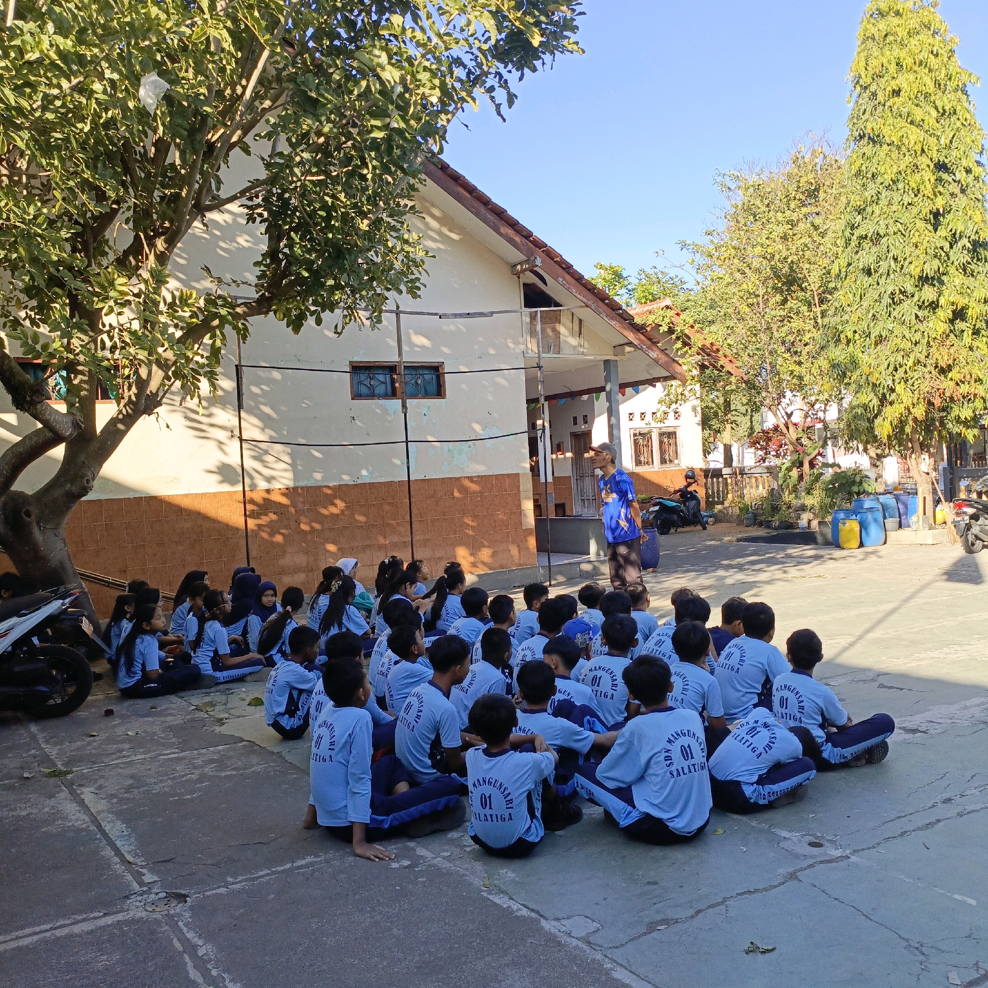 The image shows a group of young students wearing light blue t-shirts with the text "SDN MANGUNSARI 01 SALATIGA" printed on them, seated on a paved ground in front of a building. The students are mostly facing a man in a blue t-shirt and a cap, who appears to be addressing them. The building in the background is simple, with a light-colored facade and a tiled roof. Trees are present on either side of the scene, casting shadows on the ground and building. Several motorcycles are parked to the left and right of the frame, and large blue and yellow barrels are visible in the background to the right. The overall setting suggests an outdoor school activity or assembly in the city of Salatiga, Indonesia.