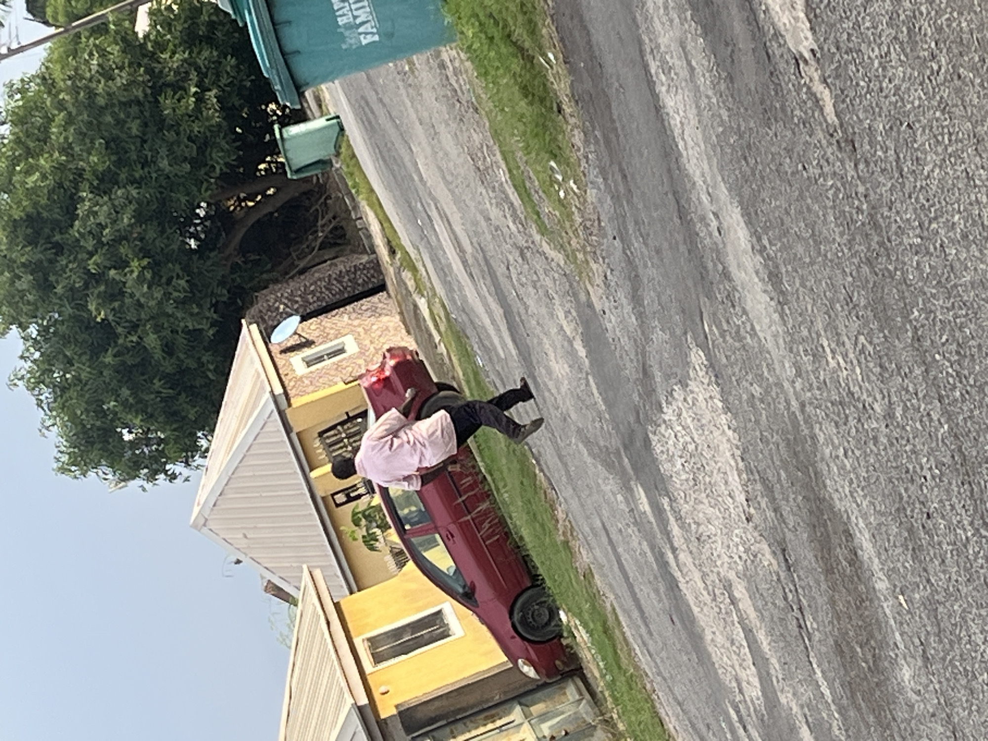 In Abuja, Nigeria, a person in a light pink shirt and dark pants walks away from a maroon sedan parked on a dirt road. The car is situated next to a yellow building with a corrugated metal roof, and a smaller stone building with a satellite dish. In the background, a large tree with dense green foliage stands tall, and two large green trash bins are visible, one with text that reads "HAPPY FAMILY". The scene is set against a pale blue sky with minimal clouds, suggesting it might be late afternoon or early morning. The ground is a mix of asphalt and dirt, with patches of dry grass.