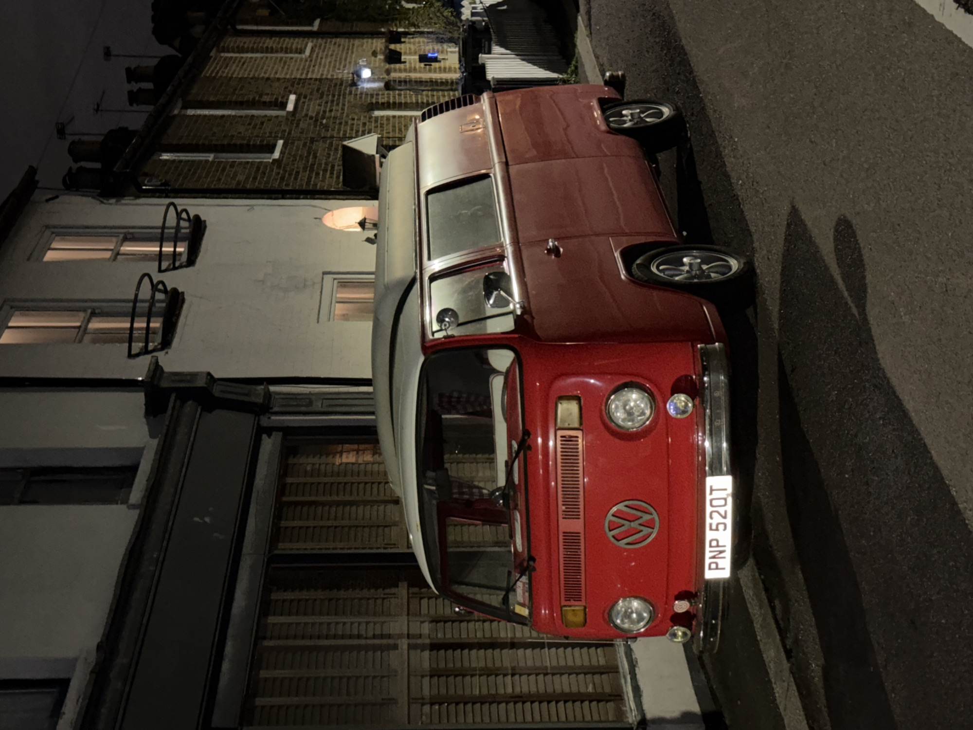 This nighttime photograph showcases a classic red and white Volkswagen Type 2 (VW Bus/Campervan) parked on an asphalt street in London, United Kingdom. The van is the prominent subject, featuring a vibrant red lower body and a contrasting white roof. Its front displays dual circular headlights, a chrome bumper, and the iconic VW emblem. A license plate, clearly visible on the front bumper, reads "PNP 52QT".
The van is positioned alongside a row of buildings. To its left, a light-colored building with multiple windows is visible, some illuminated from within, suggesting activity. These windows are adorned with decorative metal bars or brackets beneath them. Further in the background, a brick building with chimney pots on its roof also shows signs of internal lighting. Below the lighter building, a darker structure with a distinctive slatted wooden facade, possibly a large garage or shop front, is partially visible.
The scene is dimly lit by artificial sources, including internal lights from the buildings and streetlights, which cast a warm glow. A faint, elongated shadow, possibly of a person or another vehicle, stretches across the street surface to the right of the van. The overall atmosphere is quiet and still, typical of an urban street after dark. No direct human activity is depicted beyond the implied presence within the lit buildings.