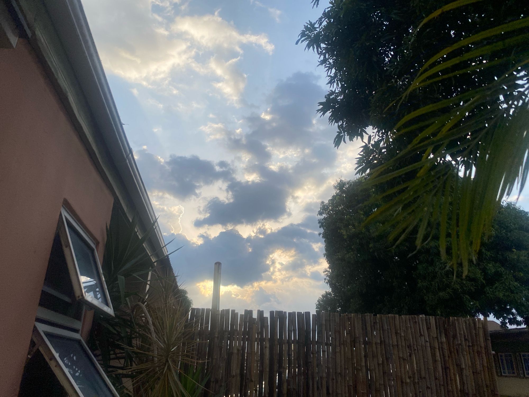 The image depicts an outdoor scene at dusk in Barden Yana, Nigeria. The sky is filled with dramatic clouds, with shafts of sunlight breaking through, creating a sense of impending or recent rain. In the foreground, the side of a pink-colored building with two open windows is visible on the left. Adjacent to the building is a tall, spiky plant. A bamboo fence runs across the mid-ground. Behind the fence, a large, dark green tree dominates the right side of the frame, with some of its leaves extending into the foreground. In the far background, a roofline of another building can be seen. A thin, vertical pipe, possibly a chimney or vent, rises from behind the fence and into the cloudy sky. There are no people or animals visible in the image.