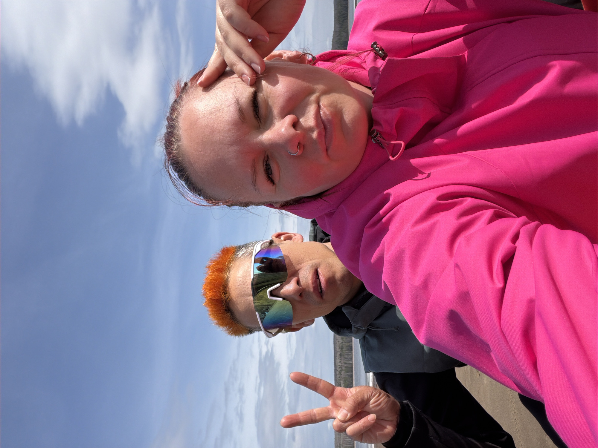 Two individuals pose for a selfie against a backdrop of sky and distant land. The person in the foreground wears a bright pink jacket. Behind this person is another individual with orange hair, sunglasses, and is making a peace sign. The location is Chaikovsky, Russia.