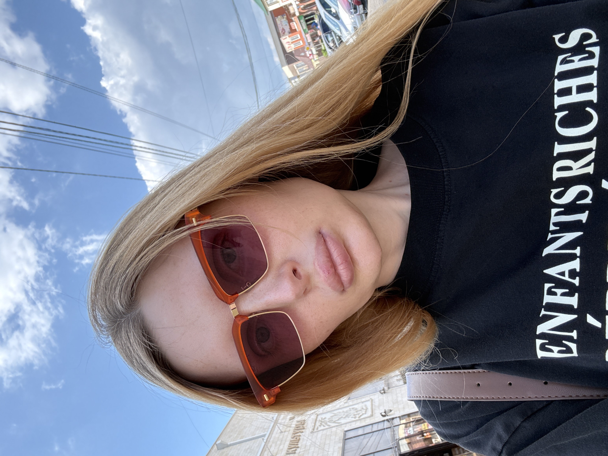 A close-up, selfie-style shot features a young woman with blonde hair, wearing large, orange-framed sunglasses with tinted lenses. She is dressed in a black crewneck sweatshirt with white text that reads "ENFANTS RICHES". The background includes a blue sky with scattered clouds, overhead power lines, and a street scene with buildings and parked cars. A portion of a building with architectural detail and text is visible on the left, and shops are visible on the right. The woman is looking directly at the camera, with her head slightly tilted. A brown strap from a bag is visible on her shoulder. The lighting suggests an outdoor daytime setting.