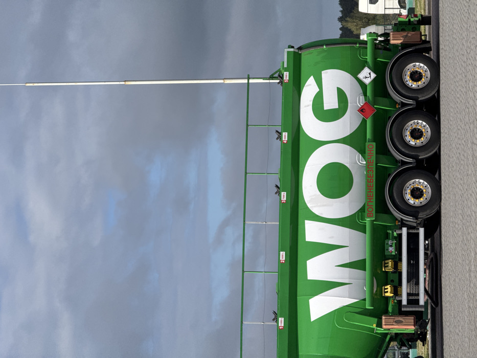 A bright green WOG fuel tanker semitrailer is parked on a light gray paved surface under a cloudy sky with visible patches of blue. The tanker prominently displays the white "WOG" logo on its side. Below the logo, a green banner with white text reads "ВОГНЕНЕБЕЗПЕЧНО" (FLAMMABLE). Further below, two diamond-shaped hazard placards are visible: a red one with a white flame symbol indicating flammability, and a black and white one with a tree and fish symbol indicating an environmental hazard. The tanker's top surface features a green metal access railing and five marked compartments, with capacities indicated as 11000 L, 8000 L, 7500 L, 5500 L, and a partially visible fifth compartment at 5500 L. A tall, slender white pole extends vertically from the forward section of the tanker, rising well above its structure into the sky. The semitrailer chassis visible features three axles with dual wheels, equipped with polished rims, black tires, and yellow lug nut indicators. Various mechanical components and a storage box are visible along the undercarriage. In the background to the right, a line of green trees and the white cabins of other commercial vehicles are partially visible. This scene is located at a commercial transport or fueling facility in Starovoitove, Ukraine.