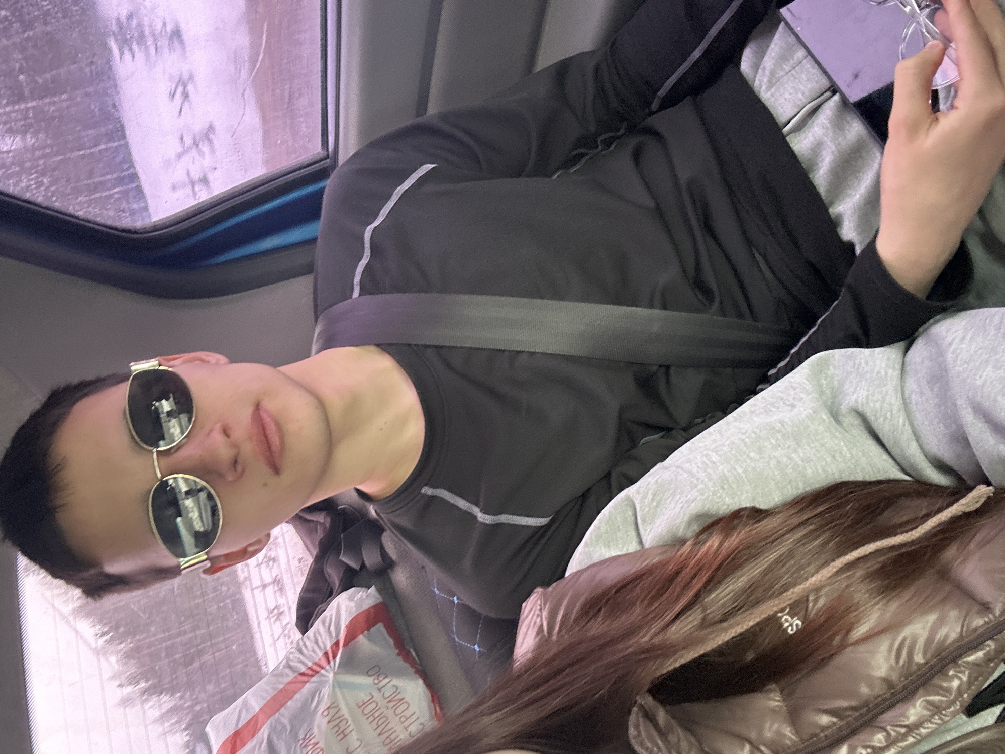 The image shows a person wearing sunglasses and a black shirt with a grey stripe, with a seatbelt across their chest, in what appears to be a vehicle. There's a window to the left. Another person's hair and part of a brown puffer jacket are also visible in the image. A package with Russian text is present. It is not possible to determine a location in the city of Ledukhi, Russia from the information given.