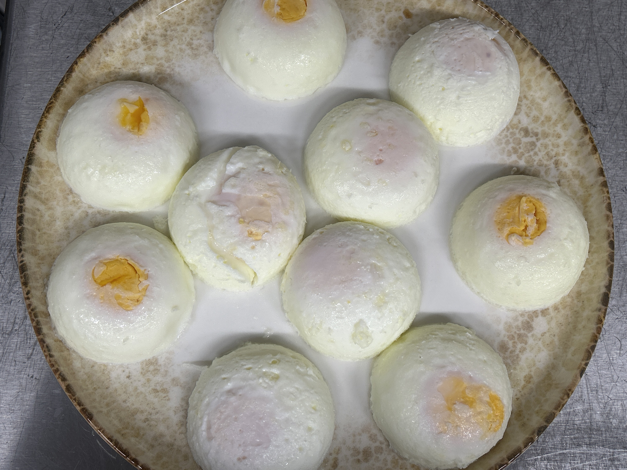 The image shows a plate of eleven poached eggs. The eggs are arranged on a speckled, shallow bowl. The background is a grey metallic surface. The scene appears to be indoors, likely in a kitchen or restaurant setting. There is no indication of the location being Burgas, Bulgaria.