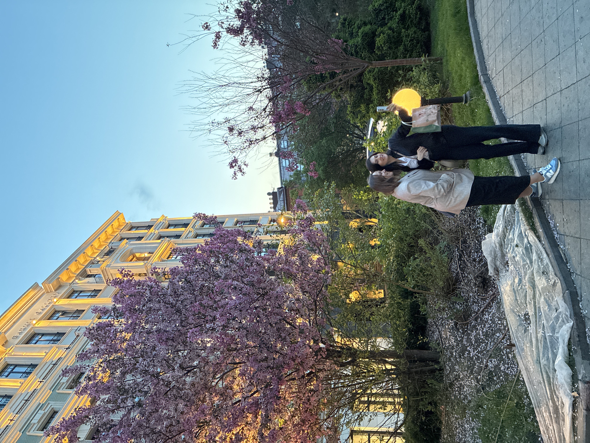 The image shows an urban scene in Moscow, Russia. Two women stand on a sidewalk next to a park-like area with trees, some in bloom with pink flowers. A building with lit windows rises in the background. One woman is wearing a light-colored jacket and a dark skirt, while the other is in a dark suit and white top. The woman in the suit holds a shopping bag and what appears to be a phone. A round lamppost provides illumination. The sidewalk is edged with a curb and has fallen flower petals scattered on the ground.