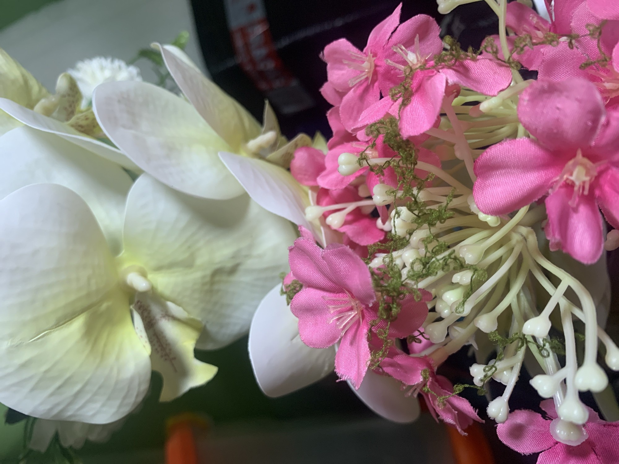 A close-up shot focuses on a bouquet of artificial flowers, dominated by vibrant pink blossoms with delicate white stamens and clusters of small, pale green buds and stems. Behind the pink flowers, larger, creamy white orchid petals with subtle yellow and brown markings are visible. The background is dark and out of focus, with a hint of an orange object on the far left and a patterned dark surface on the right, possibly a bag or packaging. The lighting is soft, highlighting the textures and colors of the floral arrangement. This image does not contain any information to determine its location in Kuala Pembuang, Indonesia.