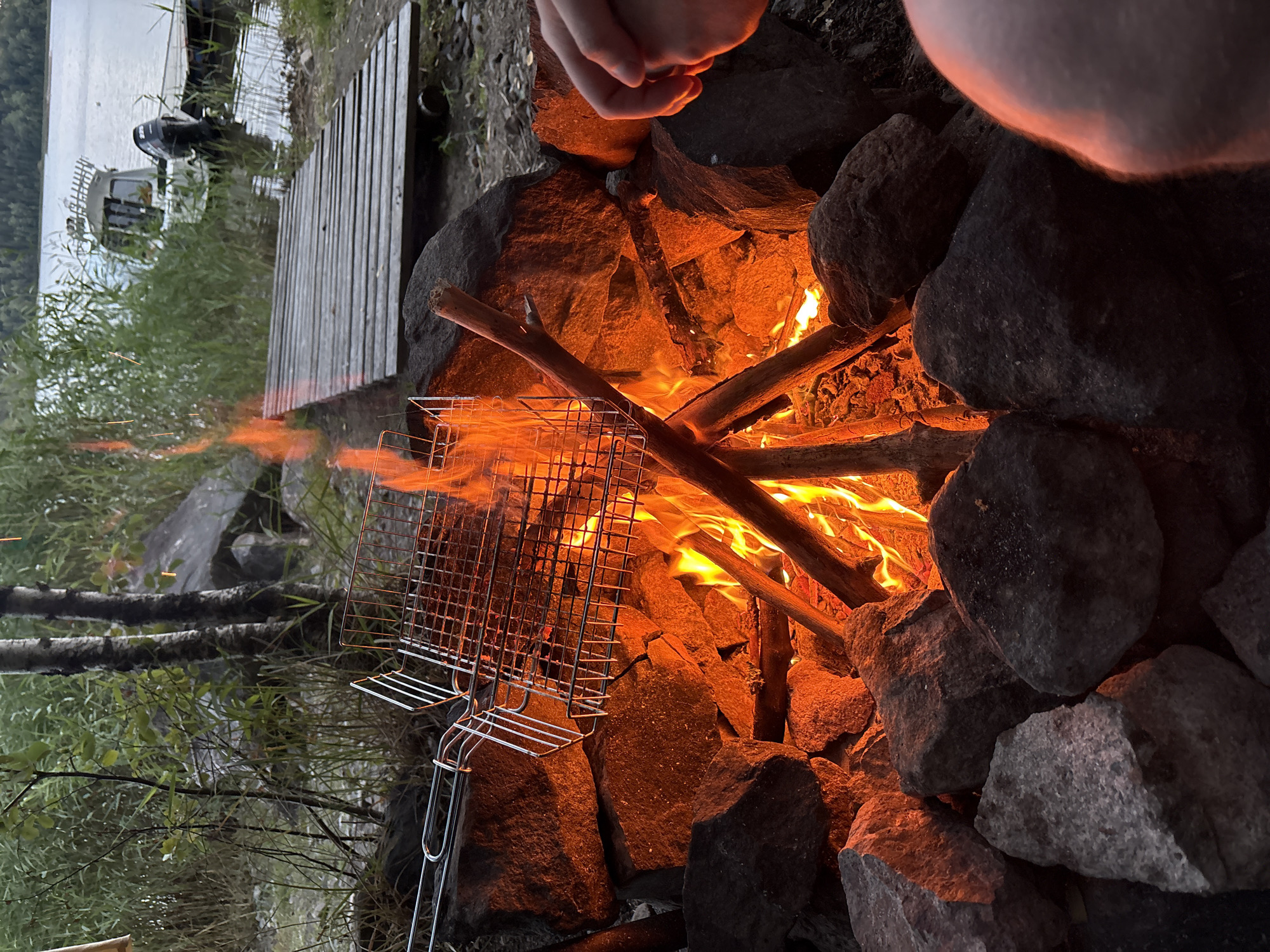 A campfire burns intensely in a rocky pit in Rautalakhti, Russia. A metal grill basket sits over the flames, indicating preparation for cooking. The surrounding area features tall grass and trees, with a wooden walkway leading towards a body of water where a boat is docked. The lighting suggests it is either dusk or dawn. A person's hand and knee are visible in the foreground, suggesting a close-up perspective of someone enjoying the campfire.