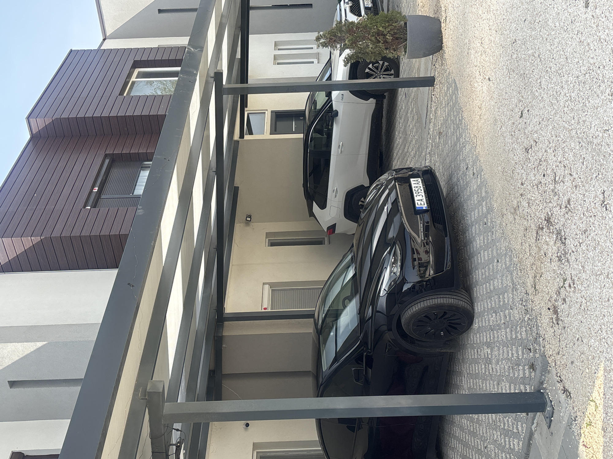 The image depicts a parking area under a metal canopy in an urban environment. Two cars are visible: a black car in the foreground with license plate "EA3958AA", and a white car behind it. Another vehicle is partially visible further back. The parking surface is paved with interlocking concrete blocks, and some gravel is present along the edge. The backdrop consists of modern buildings with light-colored facades and brown wood paneling. The location is likely Plovdiv, Bulgaria, based on the license plate format. A potted plant is situated behind the white car. The scene is static and devoid of people.