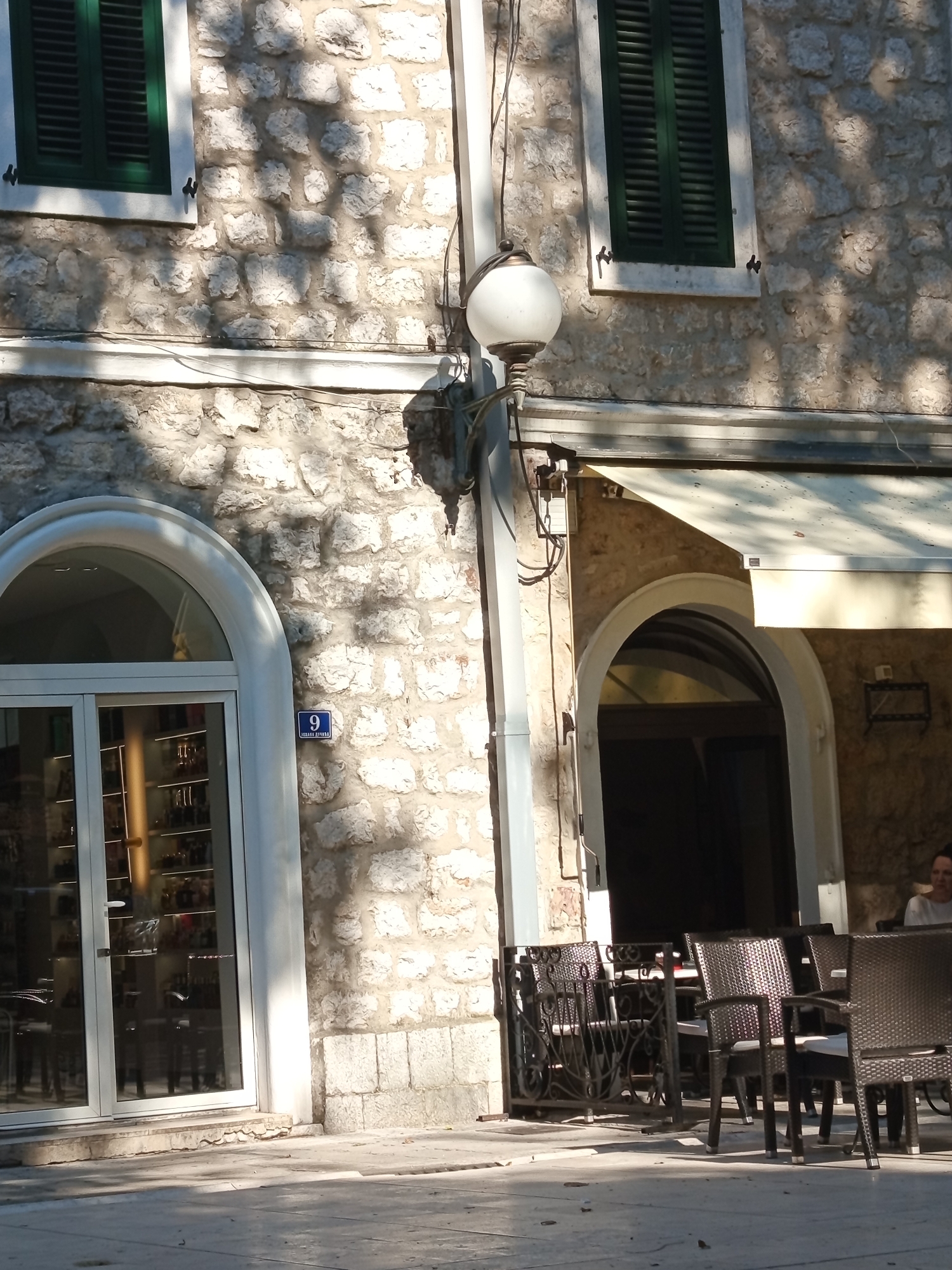 The image shows a street scene in Trebinje, Bosnia and Herzegovina. The setting is a building with a stone facade, featuring two arched entrances. The left entrance is a glass storefront, possibly a shop, with shelves of items visible inside. The right entrance appears to be the entrance to a cafe or restaurant, with outdoor seating set up. Several tables and chairs are arranged on the pavement, suggesting that patrons are expected or have recently been present.
A light fixture is mounted on the exterior wall near the left entrance, and a blue street sign with the number "9" and some unreadable text is also visible on the stone wall. Green shutters are visible on a window above the left entrance. The sun is casting dappled shadows on the stone building and the pavement, indicating it is daytime, likely mid-morning or afternoon, with clear weather. The overall atmosphere is calm and inviting, typical of a European town square or street. A person, a woman with dark hair, is partially visible at one of the outdoor tables on the right side of the image, looking to the right.