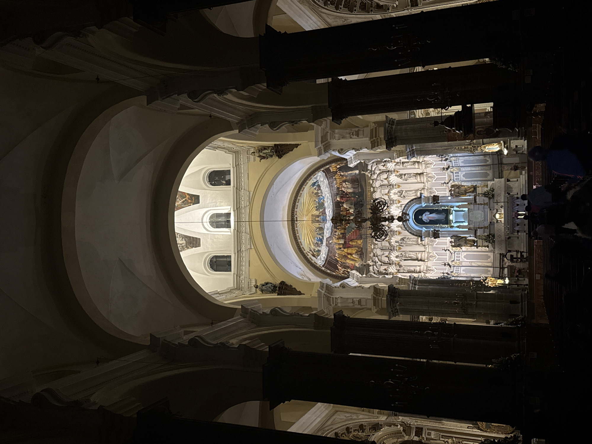 This is an interior view of a church, likely the Cathedral of Puebla, in Puebla, Mexico. The camera is oriented upward, emphasizing the high arches and architectural details. The walls are mostly white with the columns and architectural elements showing ornate decoration. The building features a semi-circular area with artwork and windows. The foreground is shadowed with columns and decorations. There appear to be people present in the shot.