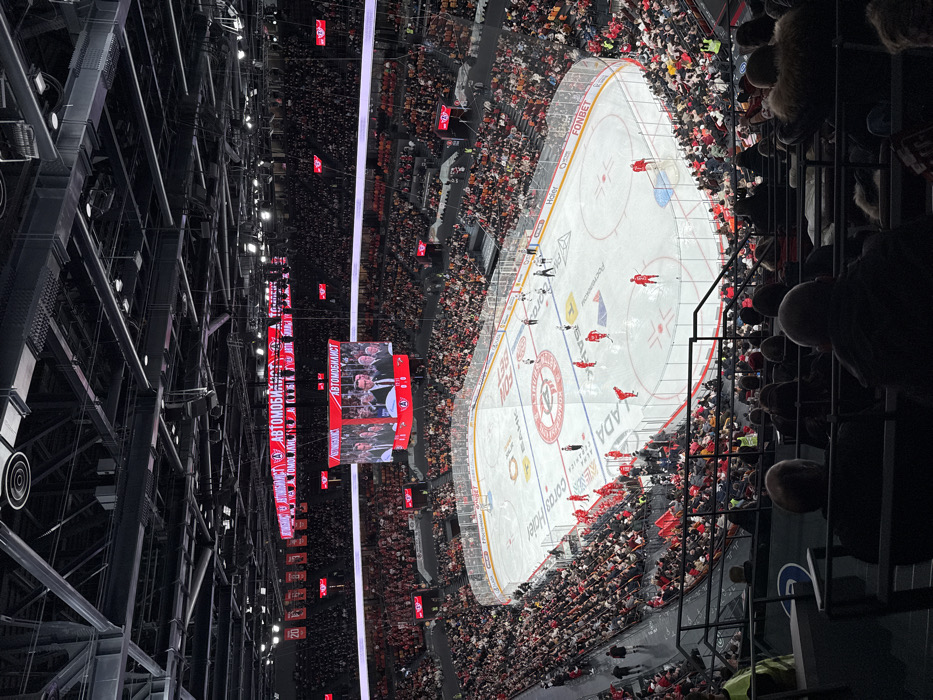 This image depicts an indoor ice hockey arena, specifically the UMMC Arena in Yekaterinburg, Russia, during an event. The scene is observed from an elevated spectator perspective, overlooking a full-sized ice rink surrounded by tiered seating filled with a large crowd.
The central ice rink features standard hockey markings, including red and blue lines, face-off circles, and two goals. Several sponsor logos are visible on the ice, including "ФОНБЕТ" (Fonbet), "КОНТИНЕНТАЛЬНАЯ ХОККЕЙНАЯ ЛИГА" (Kontinental Hockey League - KHL), "BANK", "Corsair", "LADA", and "Почта России" (Russian Post). Approximately 10-12 ice hockey players in matching red uniforms with white trim are scattered across the rink, some standing near the benches, and one positioned near a goal. Two officials in black and white striped referee uniforms are also present on the ice. A single individual in a dark suit stands near the rink barrier on the left side.
Above the ice, a large, four-sided video cube (Jumbotron) hangs, displaying a close-up image of several men in suits. The text "АВТОМОБИЛИСТ" (Avtomobilist), which is the name of the local KHL team, is prominently displayed in red and white on the video cube and on horizontal ribbon boards running along the upper perimeter of the arena. Numerous red banners with white numbers, such as "20", hang from the upper structure of the arena.
Thousands of spectators occupy the dark-colored tiered seating sections, which wrap around the entire rink. The crowd is predominantly dressed in dark attire, with some individuals wearing red. The arena's ceiling consists of a dark, exposed structural framework with various lighting fixtures. In the immediate foreground, the backs of several seated spectators are visible, positioned behind a black metal railing.