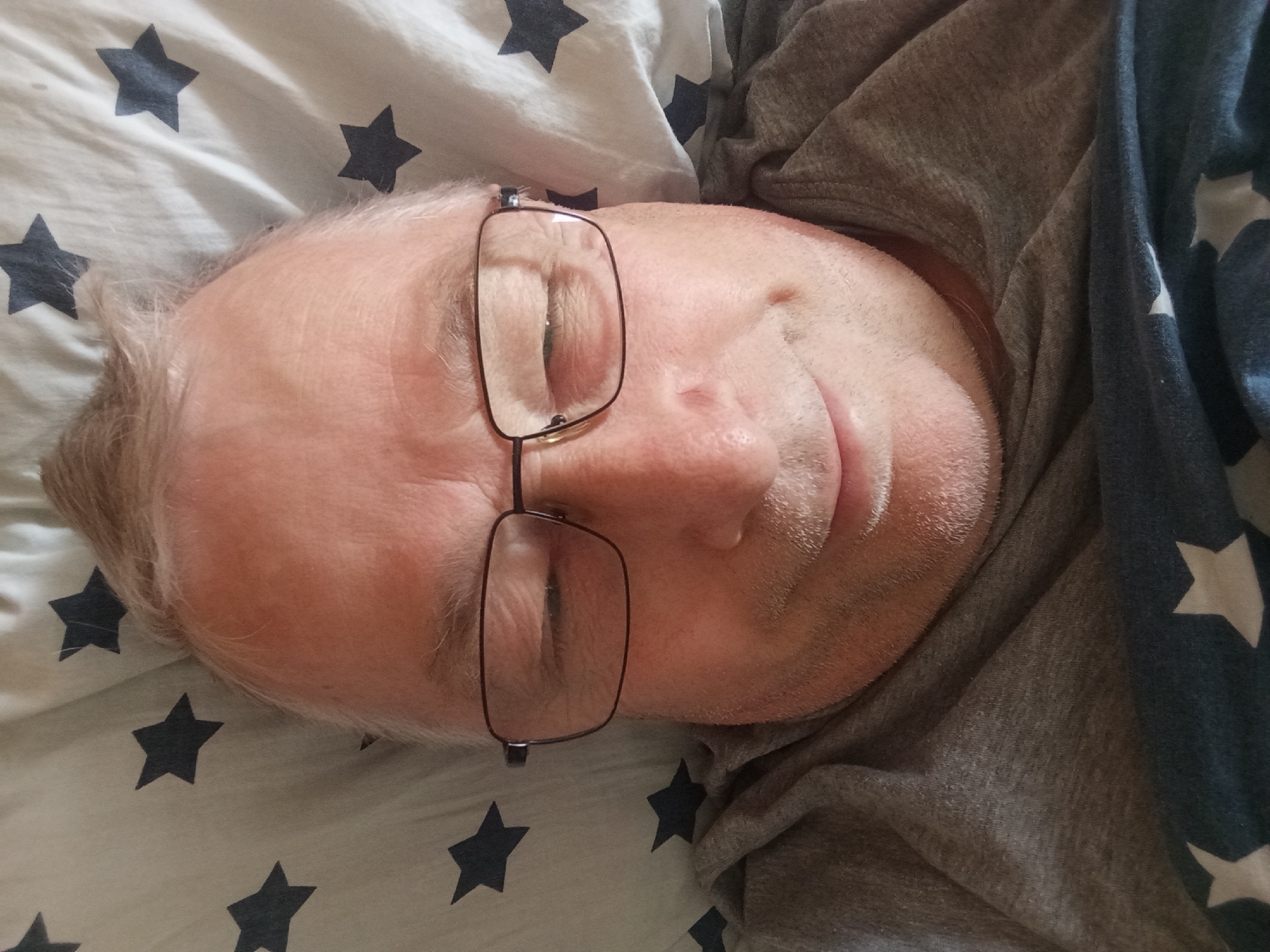 A close-up, high-angle shot captures a man resting or sleeping indoors in Nitra, Slovakia. His head, with fair skin, light-colored, thinning hair, and short facial stubble, is positioned on a light-colored duvet or sheet patterned with dark stars. He is wearing rectangular-framed glasses, which are slightly askew on his face, and a grey, casual top. His eyes are closed, suggesting peaceful sleep or deep rest. The bedding and shirt are wrinkled, indicating a relaxed, informal setting, likely a bedroom. No specific time of day or weather conditions are evident, and no text is visible.