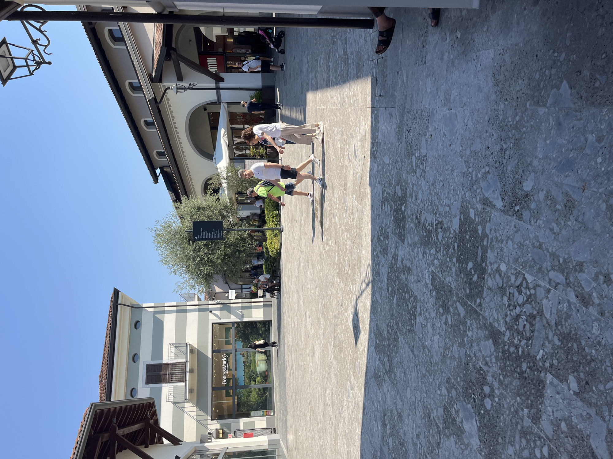 The image shows an outdoor pedestrian area in Rodengo, Italy, likely an outlet mall, on a sunny day. The ground is paved with large, light-colored stone tiles with dark speckles. In the background, there are buildings with shops, characteristic of a retail complex. One storefront displays the "Bottega Verde" logo. Several people are walking through the area. A family of four, two adults and two children, walk together in the mid-ground. Another couple with two dogs is seated on a bench to the left. A person's legs and feet are visible in the lower right corner, suggesting they are sitting or reclining. A stroller is parked near a storefront to the right. The sky is clear and blue.