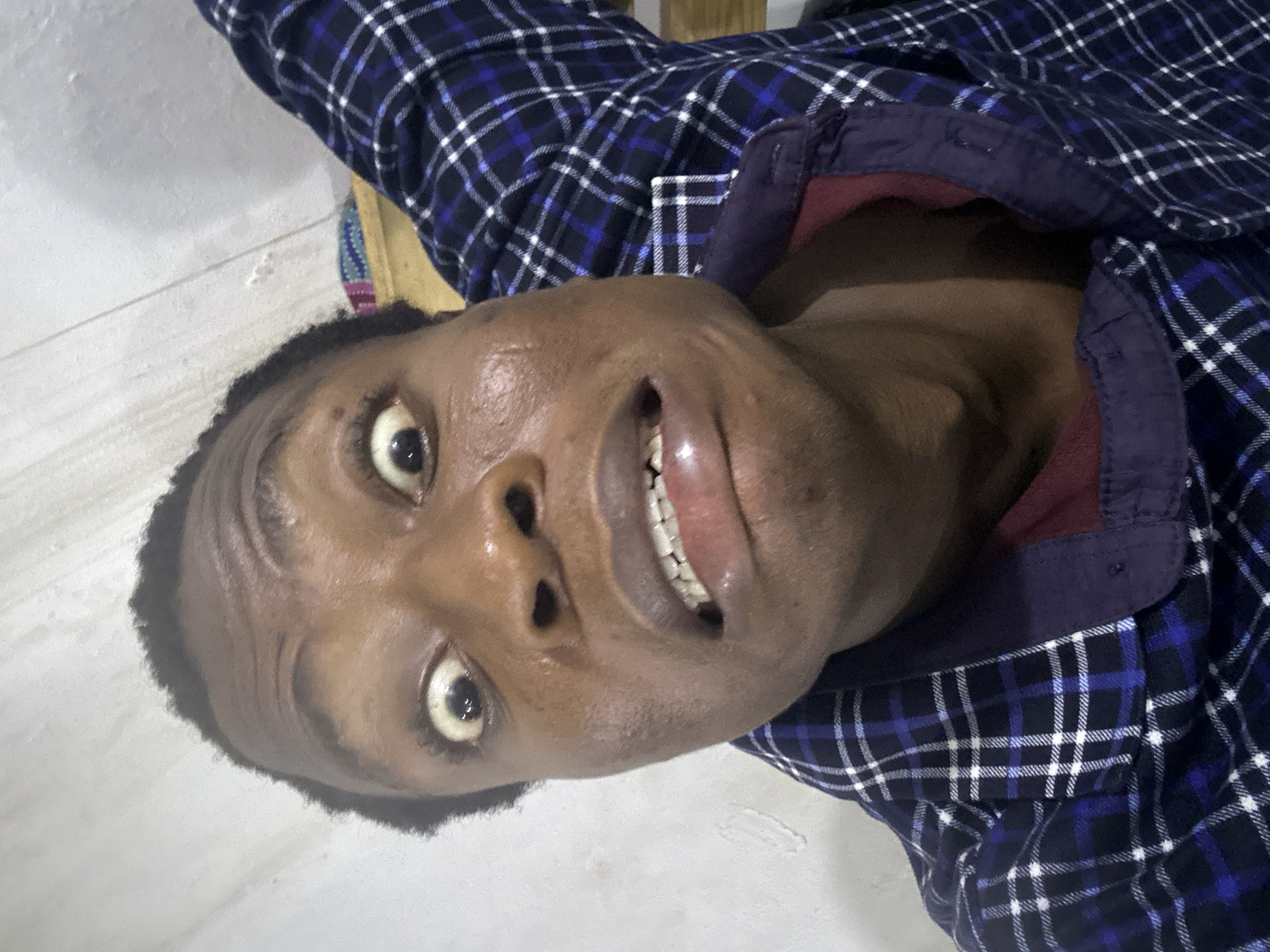 A dark-skinned person with short hair lies on a white surface. They are wearing a blue and white plaid shirt with a purple collar. The person's face is prominently featured, with visible teeth and wide eyes. The setting appears to be indoors, possibly in the city of Unknown, Unknown.