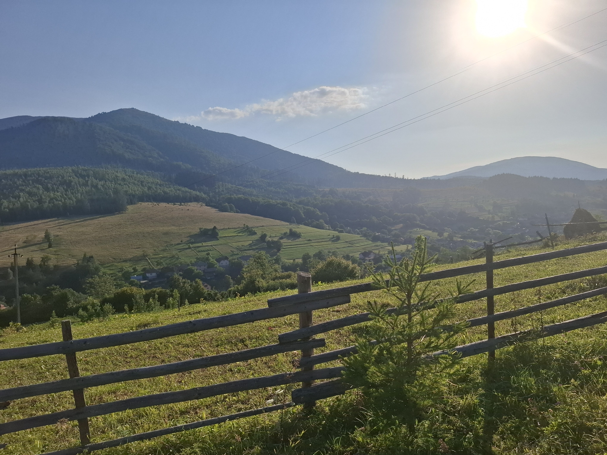 The image depicts a mountainous landscape in the vicinity of Volovets, Ukraine, under a clear sky with the sun prominently visible in the upper right corner. A rustic wooden fence made of horizontal logs supported by vertical posts runs diagonally across the foreground, separating a grassy field from a valley below. In the distance, rolling hills covered with a mix of trees and fields descend into a valley where scattered houses and agricultural terraces are visible. A few bare power lines stretch across the sky. The overall scene conveys a serene, rural, and natural environment. There are no people or animals present.