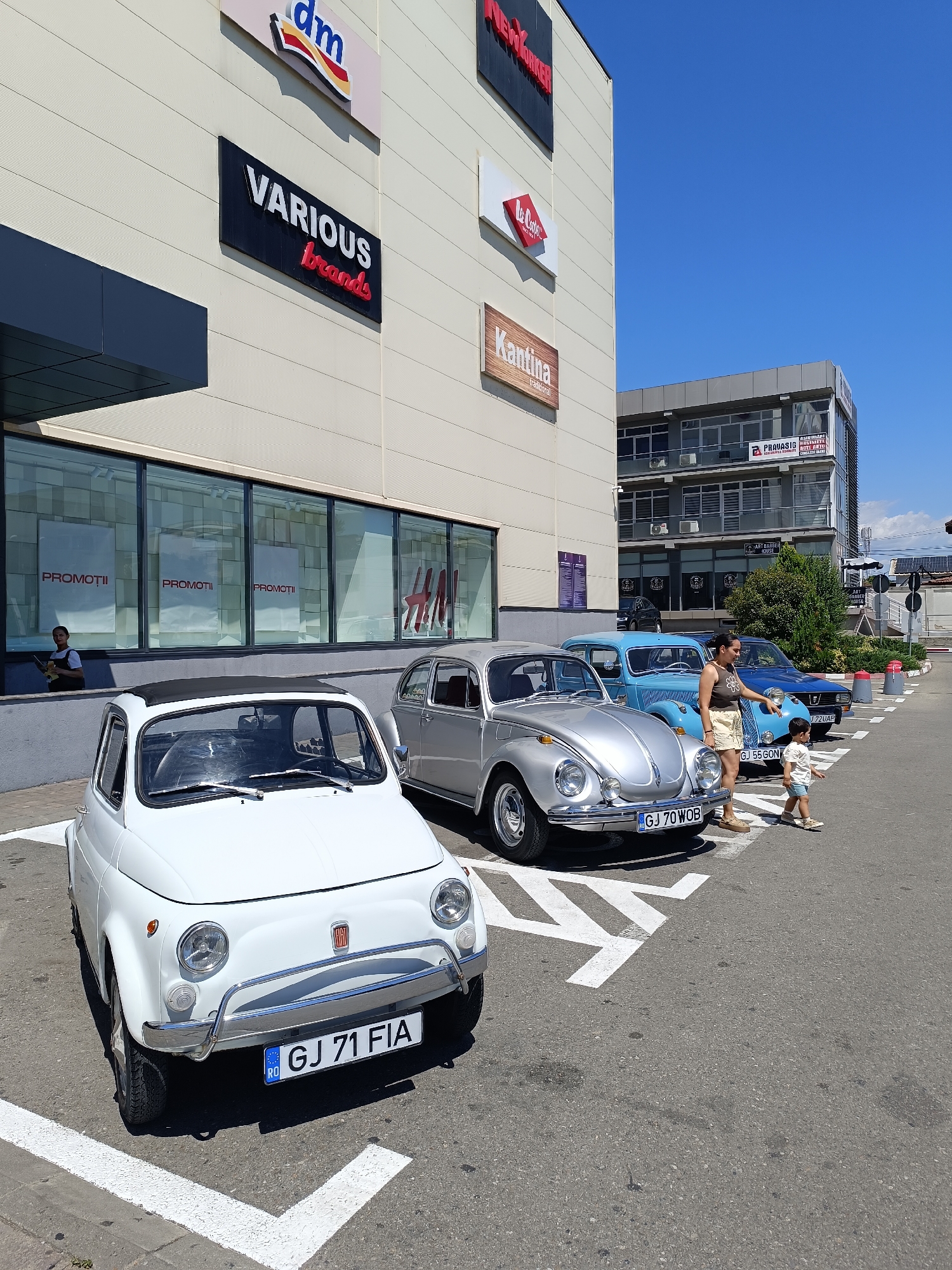 In Târgu Jiu, Romania, a white Fiat 500, a silver Volkswagen Beetle, and two blue cars are parked in a lot in front of a modern building with storefronts. The building features signage for "dm," "VARIOUS brands," "NewYorker," "La Co.,." and "Kantina." In the foreground, the Fiat 500 has a license plate reading "GJ 71 FIA." The silver Volkswagen Beetle has a license plate "GJ 70 WOB." A woman and a young child are standing near the Volkswagen Beetle, with the woman pointing towards the blue cars. Behind the parked cars, a multi-story building with offices is visible, displaying a sign for "PRAVASIG." The sky is clear and blue, indicating a sunny day.
