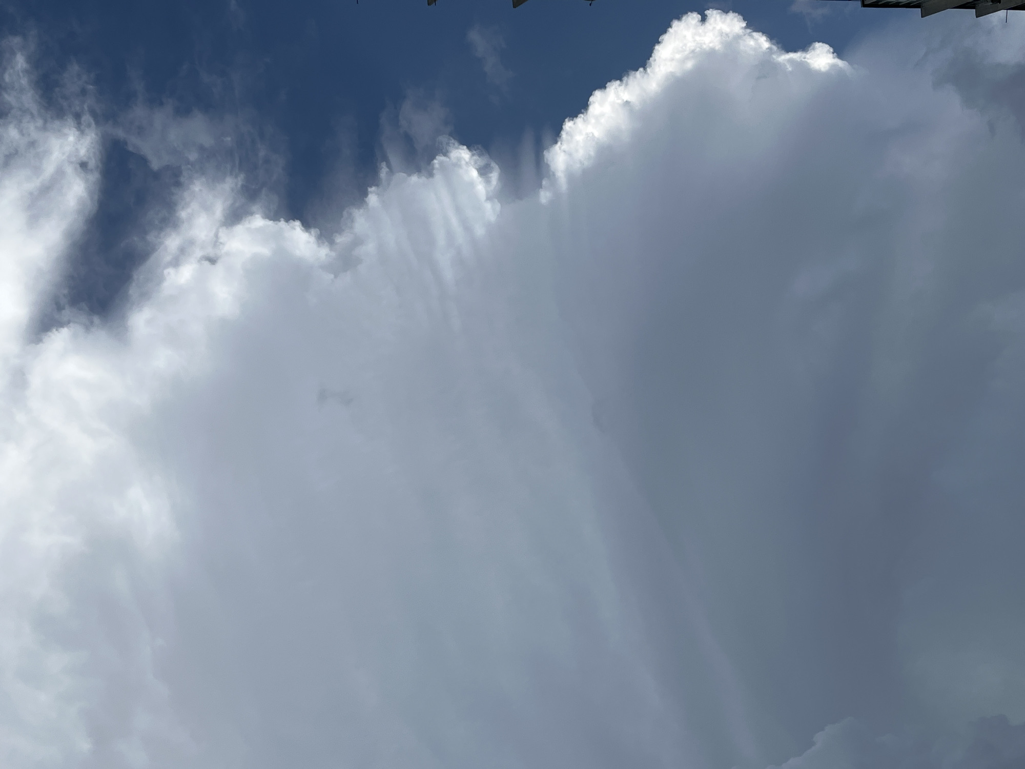 An outdoor, vertical, medium shot captures a large, grey cloud formation dominating the left and center of the frame, with a sliver of bright blue sky visible on the right. The cloud exhibits a dramatic, jagged edge where it meets the sky. Wispy, lighter clouds are layered on top of the main cloud formation, creating a sense of depth and texture. The bottom right of the image shows the top of a dark, angular building structure, with its details obscured by the lighting. The overall lighting suggests it is daytime, with the sun illuminating the edge of the clouds. The location is identified as Unknown, Unknown.