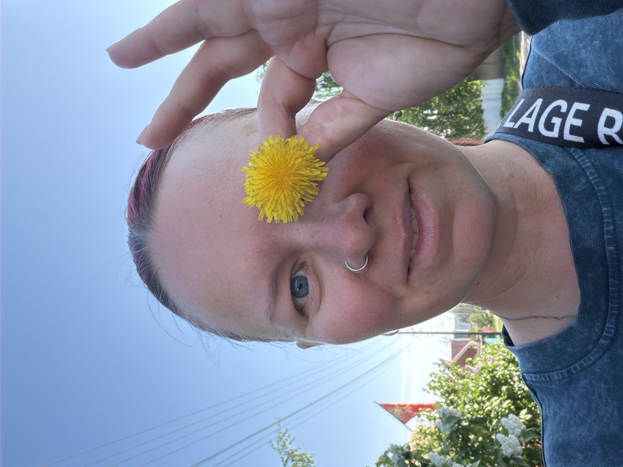 A woman is holding a yellow dandelion flower in front of her face. The flower is positioned over her nose. She has fair skin, light hair with pink highlights, and a silver nose ring. She is wearing a blue denim shirt with a black strap that reads "LAGE R" visible. The background consists of a blue sky, power lines, greenery, and a partially visible building. The location is potentially Chaikovsky, Russia.