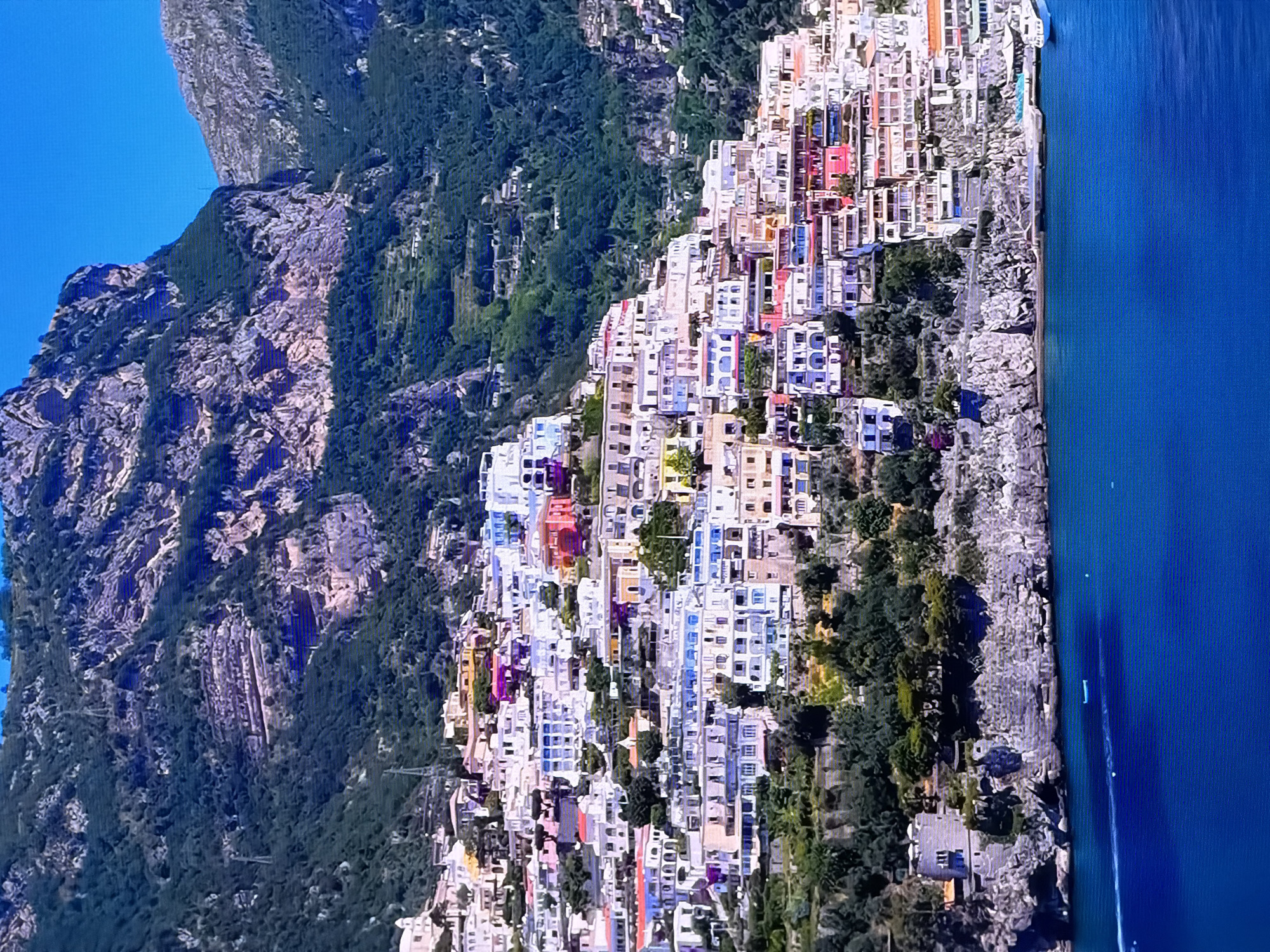 The image displays a panoramic view of a colorful, densely packed town built on steep cliffs overlooking a vibrant blue body of water. The architecture is characterized by buildings with white, pastel, and brightly colored facades, stacked closely together and cascading down the mountainside. Many structures feature balconies and tiered levels. Lush green vegetation, including trees and shrubs, is interspersed throughout the town and covers the surrounding steep, rocky mountains. The water in the foreground reflects the bright blue sky, and a white boat is visible on the surface. The scene is bathed in natural sunlight, creating stark contrasts and highlighting the textures of the buildings and the natural landscape. There are no people visible in the image.
The provided image does not depict the city of Chirchik, Uzbekistan. The scene is characteristic of coastal towns in Mediterranean regions, such as Positano on the Amalfi Coast in Italy, known for its dramatic cliffside villages.