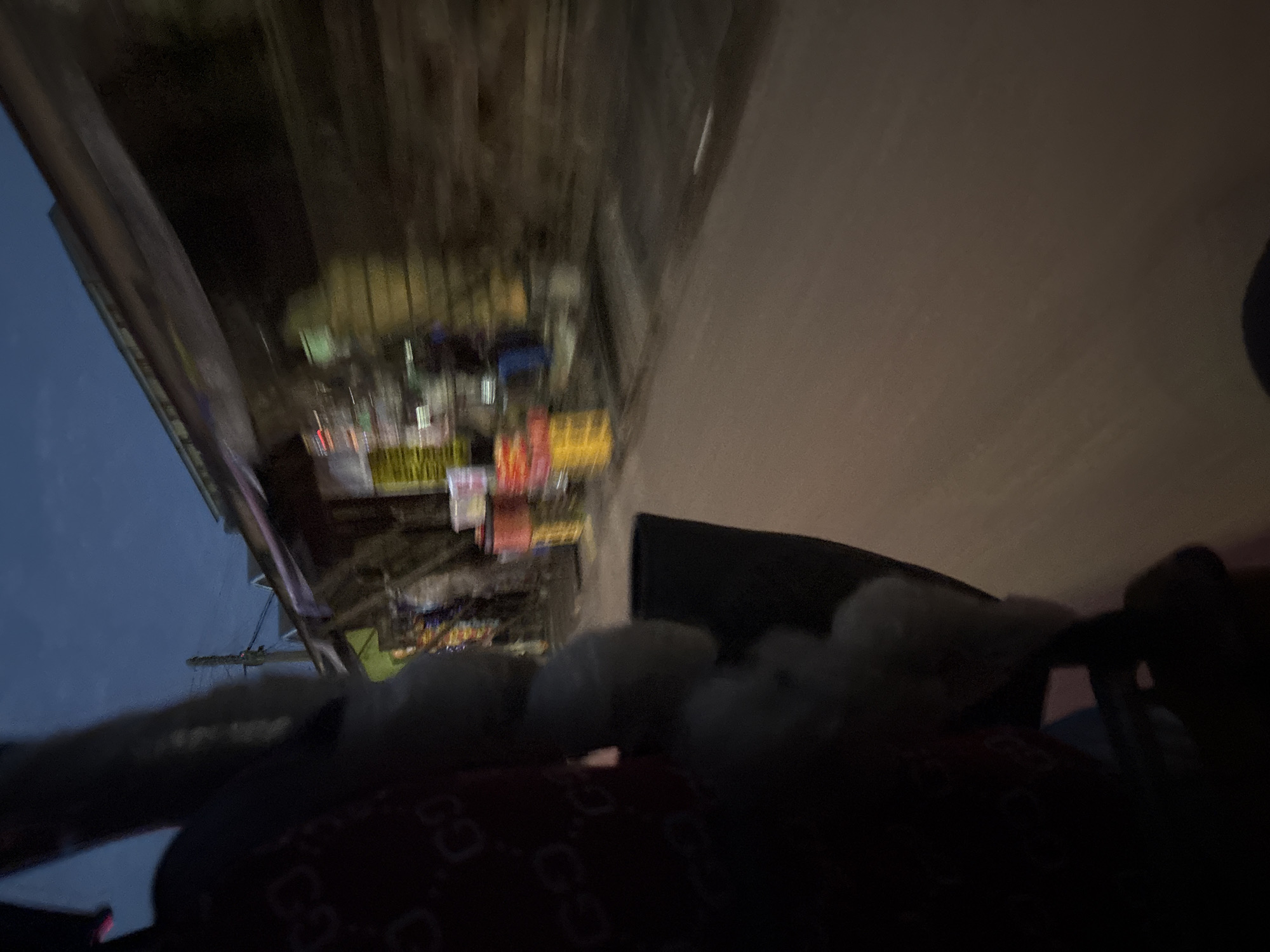 The image depicts a street scene at dusk or night in Onitsha, Nigeria. In the foreground, a portion of a person's dark clothing, possibly a shirt with a repeating pattern, is visible on the left. Next to it, a black bag or item is positioned. The background shows a blur of activity along a street lined with shops or stalls. Various colorful items, likely goods for sale, are displayed on shelves or carts. The sky above is a deep blue, indicating the late hour. The motion blur suggests the image was taken from a moving vehicle. The overall atmosphere is bustling, typical of a market area in a Nigerian city at night.
