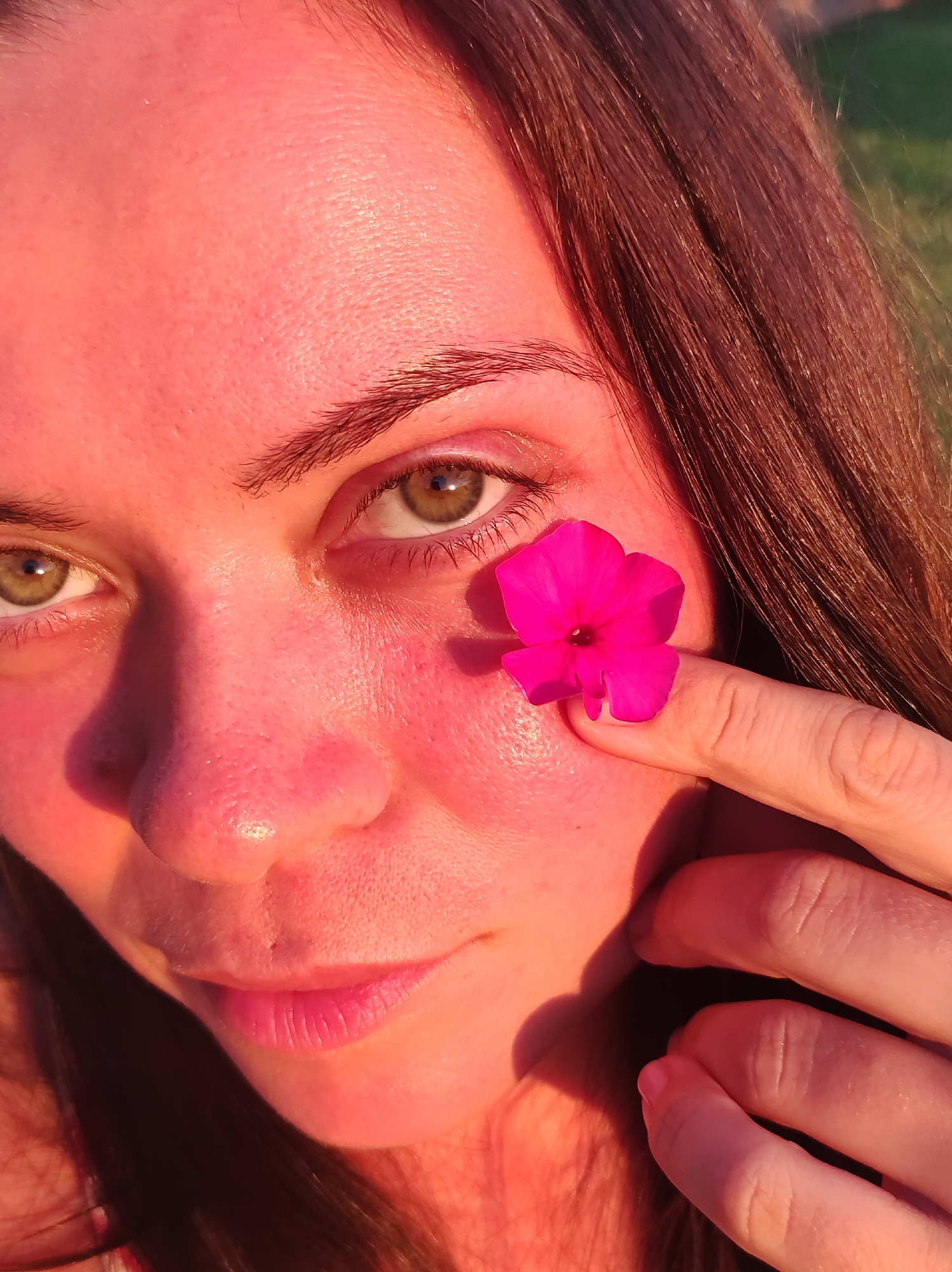 The image is a close-up, eye-level portrait of a young woman with brown hair, outdoors during the golden hour. She is looking towards the camera with a slight smile. A bright pink flower is held on her cheek by her right index finger, which is positioned just below her right eye. Her skin is flushed pink from the warm sunlight, highlighting texture and pores. Her eyes are green and her eyebrows are well-defined. The background is blurred, suggesting a grassy area bathed in sunlight. The overall lighting is warm and soft. There is no identifiable location within the city of Фоки, Russia, visible in this image.