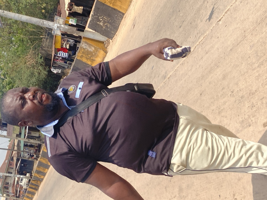 A male individual with a dark complexion, bearded, and of stout build, is captured mid-stride on a paved road in Afikpo, Nigeria. He wears a dark brown polo shirt featuring a light blue collar, placket, and a gold-colored "B" emblem on the left chest, with a small white tag reading "YOU" on the lower left torso. His lower attire consists of light beige trousers with a dark stripe along the side. A black messenger bag is slung across his left shoulder. In his right hand, he grips a folded, blue and white checkered cloth. His gaze is directed forward and slightly downward, with his mouth slightly ajar.
The environment indicates a bustling street scene. To the subject's right, a paved road surface shows cracks and seams, bordered by yellow and black striped concrete barriers. Beyond these barriers, several makeshift stalls or small shops are visible, displaying clothing and featuring corrugated metal roofs; one stall has a partially visible sign indicating "SANITARY STORE". A light grey utility pole stands upright near the barriers. In the deeper background on the right, additional indistinct figures are present, one appearing to carry an item on their head. To the subject's left, multi-story buildings, possibly residential or commercial, line the street, with a white vehicle partially visible near one structure. Lush green foliage and trees are present further in the background. The scene is illuminated by bright daylight, casting distinct shadows.