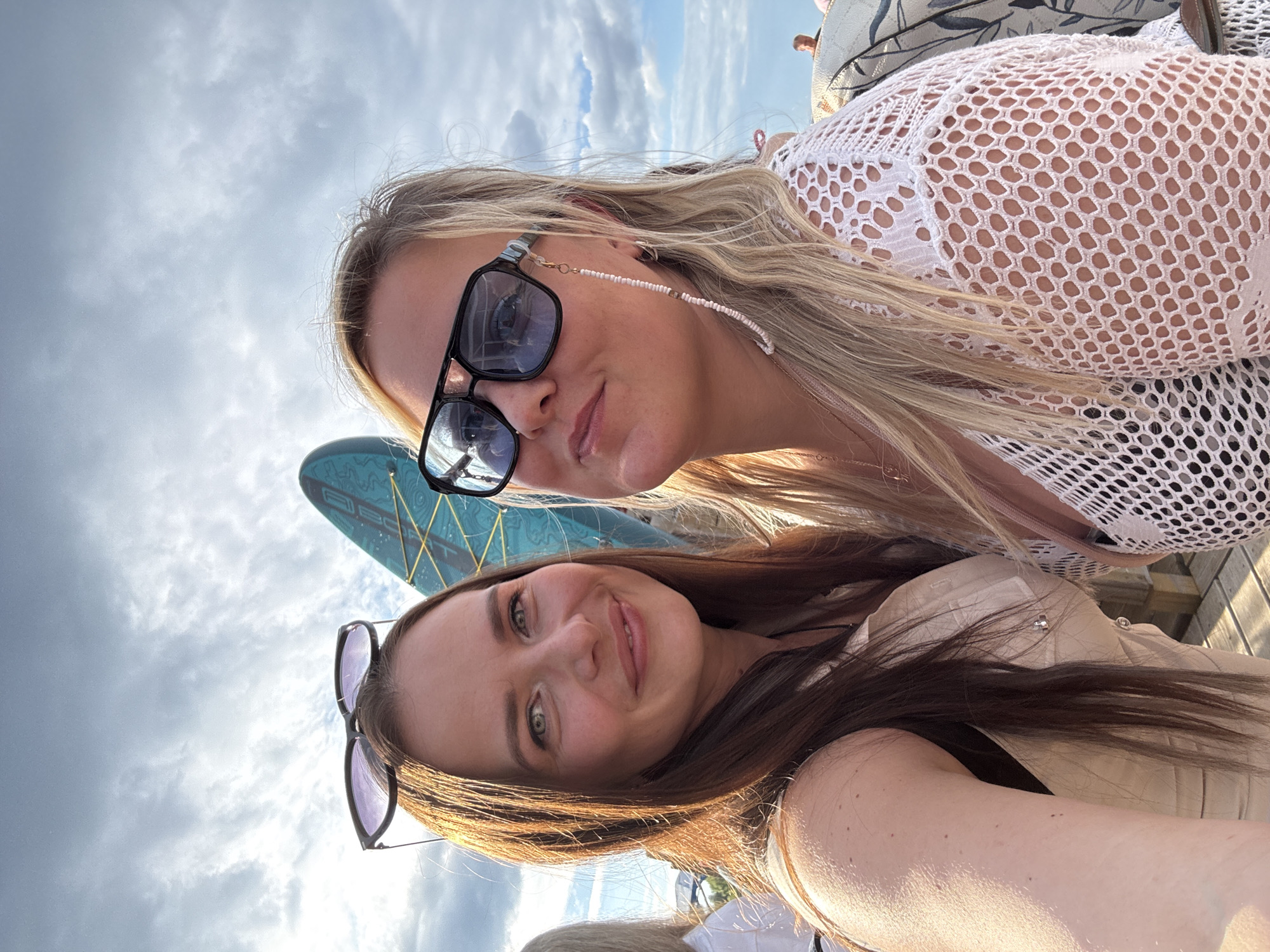 The image shows two women, likely in their 20s or 30s, taking a selfie outdoors. The woman on the left has long, dark brown hair and is wearing a light-colored top. She is smiling and looking towards the camera. The woman on the right has blonde hair and is wearing large, dark-rimmed sunglasses and a white, open-knit top. She is also looking towards the camera, with a slight smile.
Behind them, a bright blue surfboard is visible, leaning against a structure. The sky is partially cloudy, with a mix of blue sky and white clouds. The lighting suggests it is daytime, possibly late afternoon due to the warm tones on the left side of the image. The overall setting appears to be an outdoor recreational area, possibly near a body of water, given the surfboard. The bottom part of the image shows wooden planks, suggesting a deck or pier.
While the image itself does not provide direct evidence of the location being Chaikovsky, Russia, it is important to note that Chaikovsky is known for its recreational opportunities, particularly related to water sports and outdoor activities. The presence of a surfboard aligns with such potential activities. However, without further context or landmarks, pinpointing the exact location within Chaikovsky is not possible based solely on this image.