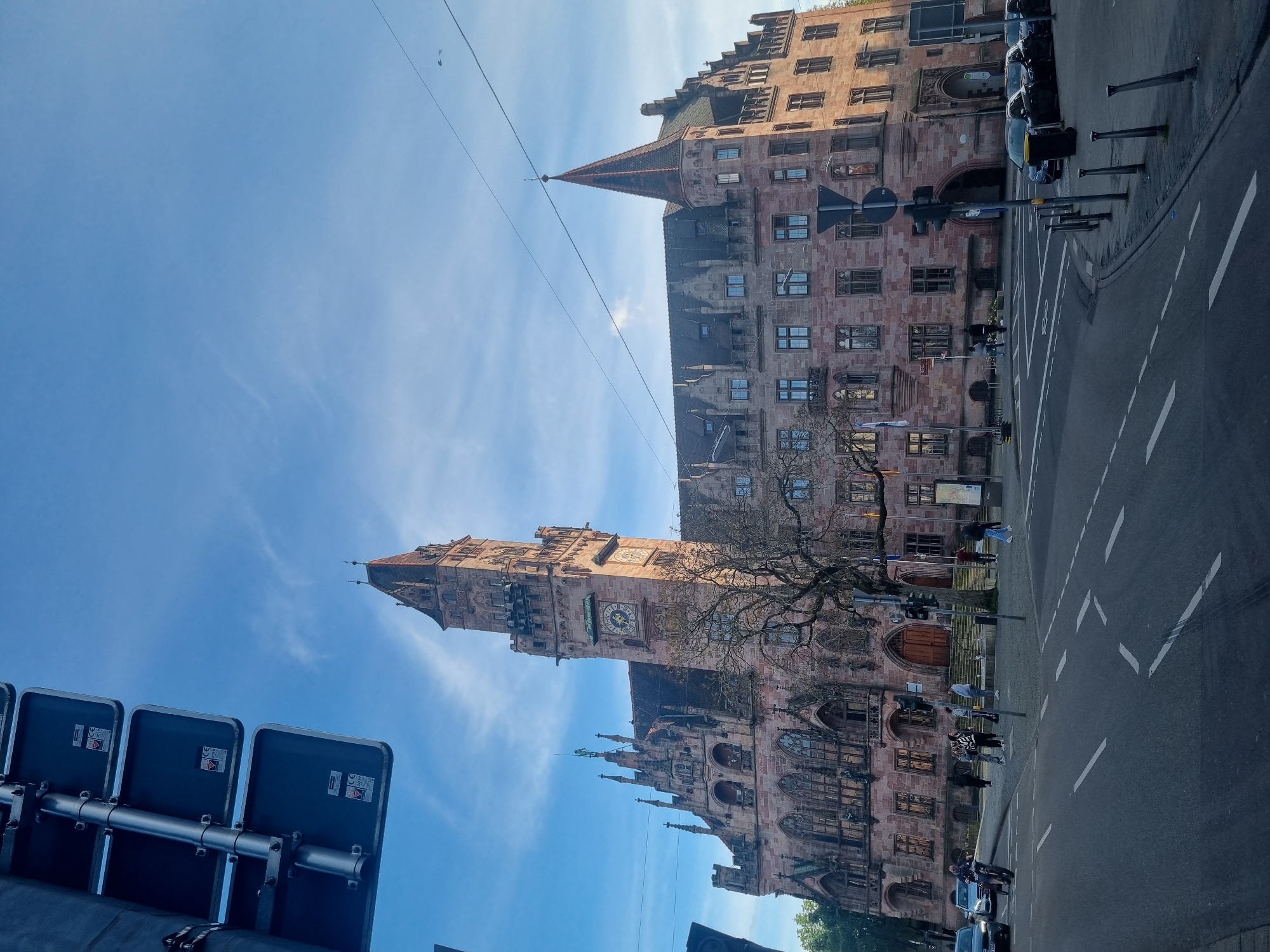 The image depicts a tall, ornate, brownstone building with multiple towers and spires, situated on a city street. The building is likely a historical landmark, given its architectural style. A clock is visible on one of the towers. Bare tree branches partially obscure the building's facade.
A street runs along the right side of the frame with white lane markings and parked cars are present. Traffic lights and signs are installed along the curb.
Several people are walking on the sidewalk in front of the building and crossing the street.
The sky is blue with a few wispy clouds.
The location is not Saarbrücken, Germany. The building is the Nuremberg Town Hall in Nuremberg, Germany.
