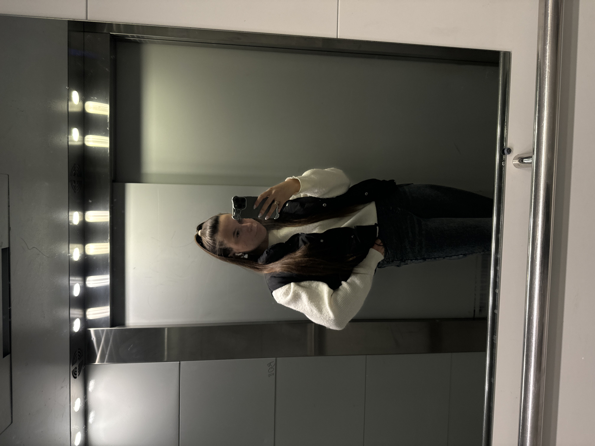 A young woman is seen in a mirror selfie. She is holding a smartphone and looking at its screen. The setting appears to be an elevator in what seems to be an apartment building or business complex, based on the interior design and lighting. The elevator has integrated lighting along the side and panelled walls. The mirror is bordered by a white frame. The location is implied to be in Odintsovo, Russia.