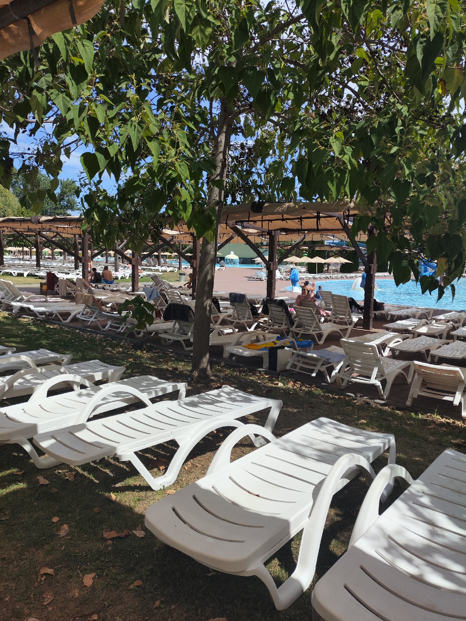 The image depicts an outdoor seating area at a water park, likely located in Sociteni, Moldova. Rows of empty white plastic lounge chairs are set up on a grassy area under the shade of trees and wooden canopies. In the background, a swimming pool with bright blue water is visible, with some lounge chairs and umbrellas arranged around its perimeter. Several people are seated on lounge chairs in the background, some appearing to be relaxing or talking. The overall scene suggests a leisure environment on a sunny day.