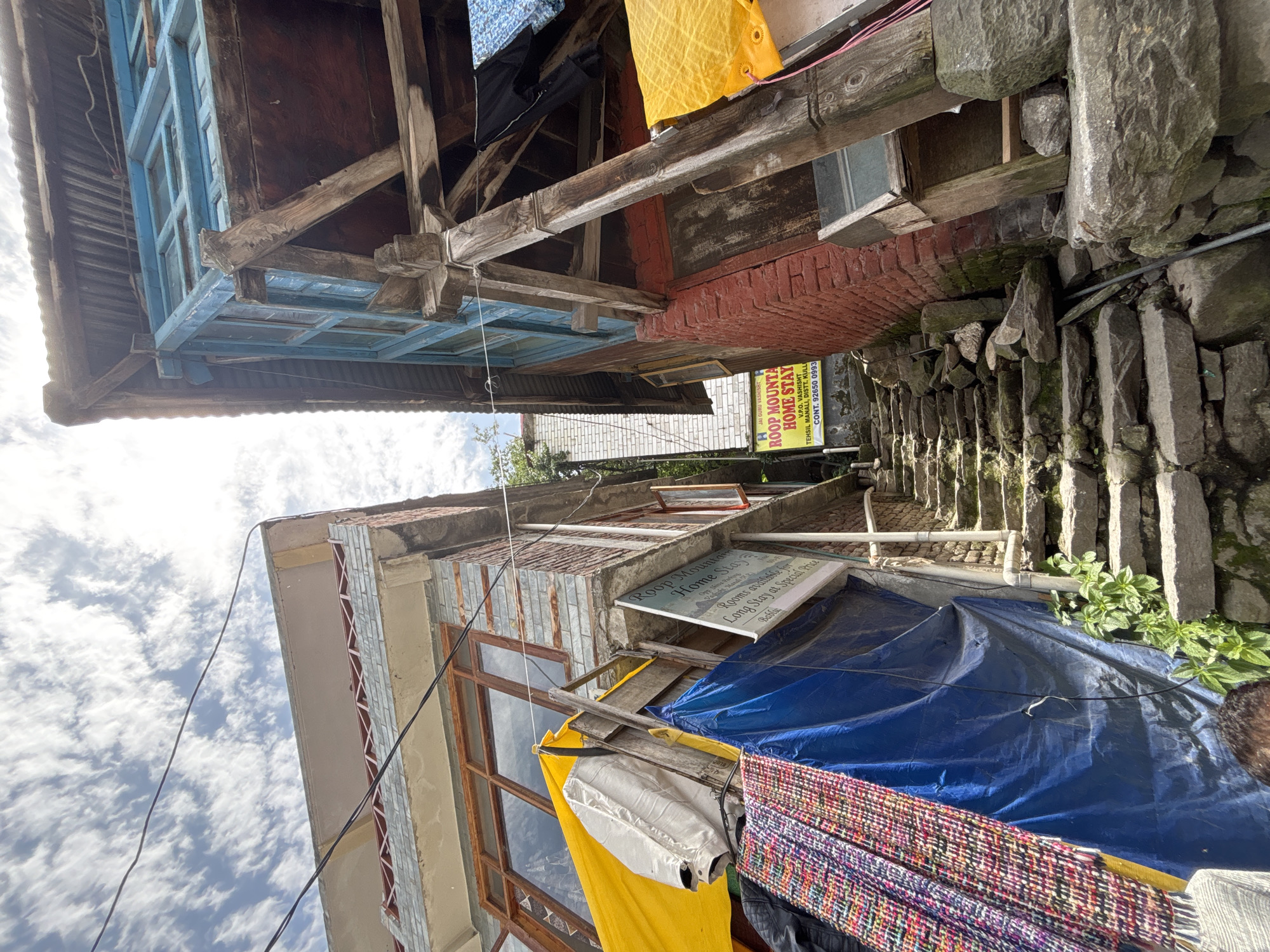 This image depicts a narrow, stone staircase in the city of Vashisht, India, flanked by two buildings. The building on the left is constructed with a grey brick facade and features a wooden-framed window. A yellow tarp is draped over a wooden structure on the left side of the staircase, partially obscuring the building. Below the tarp, there are stacks of colorful textiles. A blue tarp is also visible, hanging behind the textiles. A white sign with red lettering is attached to the left building, advertising "ROOP MOUNTAIN HOME STAY" with contact information.
The building on the right is made of red brick and has a blue wooden balcony with multiple window panes. Wooden support beams are visible, extending from this building and supporting a corrugated metal roof. Black and blue clothing is hanging to dry on a clothesline attached to the right building. The stone staircase itself is uneven and shows signs of age and wear, with moss growing on some of the stones. Lush green plants are growing along the base of the stairs and on the surrounding walls. The sky above is partly cloudy, with white clouds against a bright sky. The scene appears to be a street or alleyway, typical of older settlements in India.