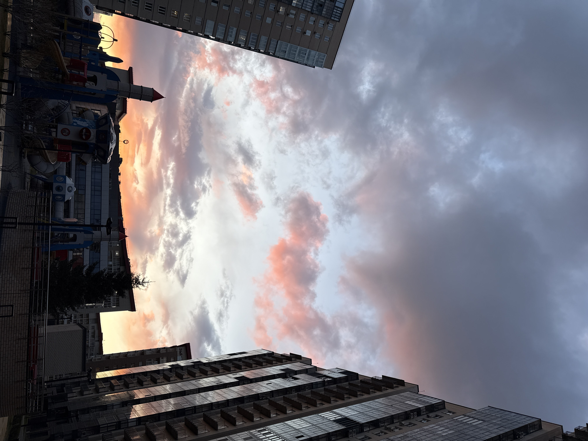 The image, taken from a low angle, depicts a cityscape in Krasnoyarsk, Russia, during a sunset. The sky is filled with gray and pink clouds, creating a dramatic effect. Two tall buildings frame the top and bottom of the image. The lower building features glass-paneled balconies. A playground structure, including a rocket ship, is visible to the left. The sun is reflecting on the side of some buildings. A tree is also visible.