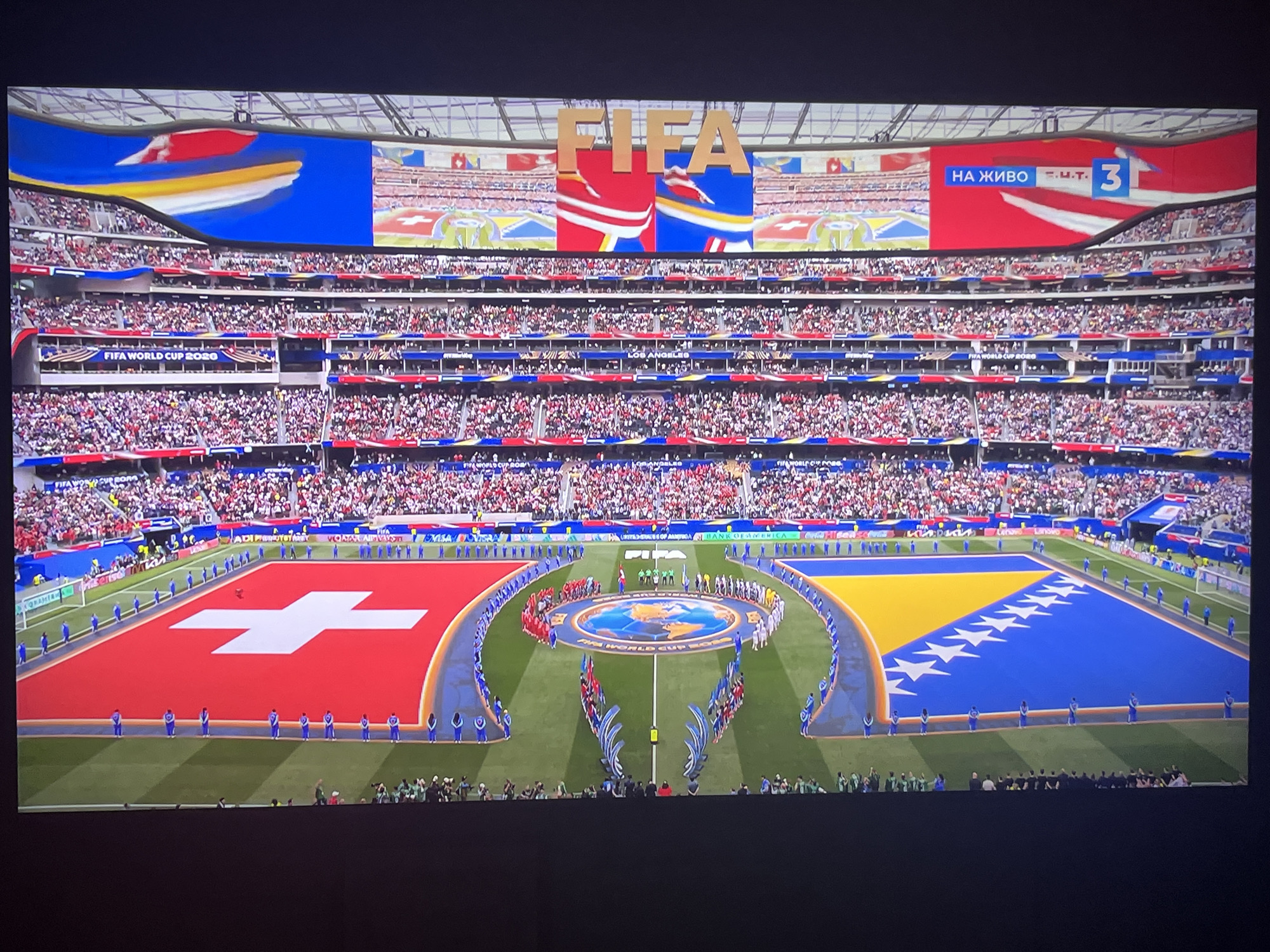 The image shows a wide-angle view of a stadium during a FIFA World Cup event. Two large national flags, the Swiss flag and the flag of Bosnia and Herzegovina, are displayed prominently on the field, covering a significant portion of the playing surface. Players and officials are lined up on the field between the flags, forming two lines that converge at the center circle. The center circle itself features a graphic of the Earth. The stadium is filled with a large crowd of spectators in the stands, many of whom are wearing red and white or red and blue attire, suggesting support for one of the teams. Large screens are visible above the stands, displaying the FIFA logo and other graphics, including text in Russian that reads "НА ЖИВО" (live). There are also advertisements visible in the stands. The image appears to be taken from a screen, possibly a television or a large display, showing the event. The specific location in Varna, Bulgaria, is not depicted in the image itself.
