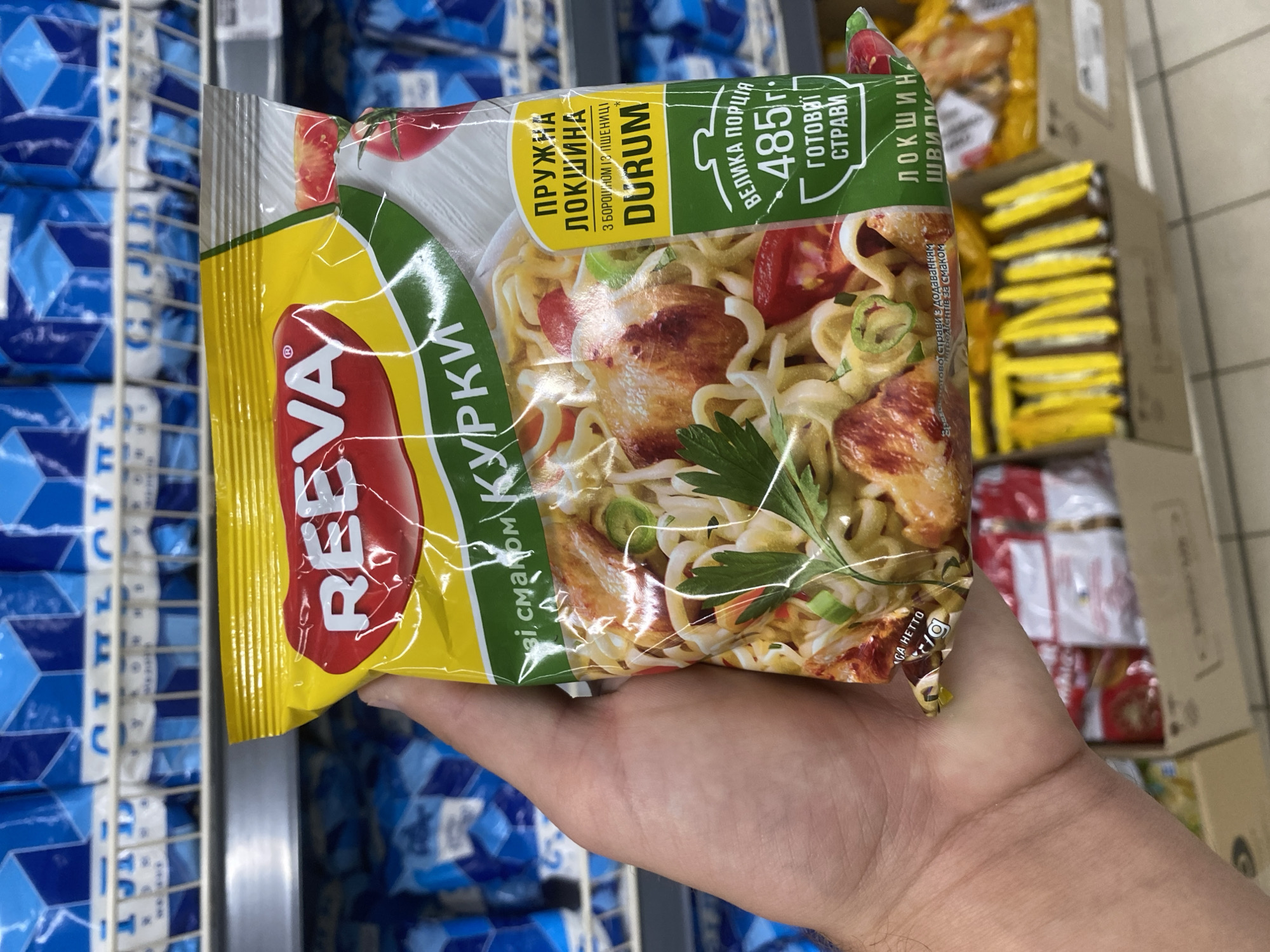 A person's hand holds a package of REEVA brand instant noodles with chicken flavor. The package is yellow and green with images of noodles, chicken, tomatoes, and green onions. Text on the package is in Ukrainian and includes "ПРЯЖНА ЛОКШИНА" (springy noodles) and "485 г ГОТОВЇ СТРАВИ" (485g ready meal). The noodles are described as being made from wheat. The background shows shelves stocked with similar blue packaged products, indicating a retail environment, likely a supermarket. The scene is indoors.
