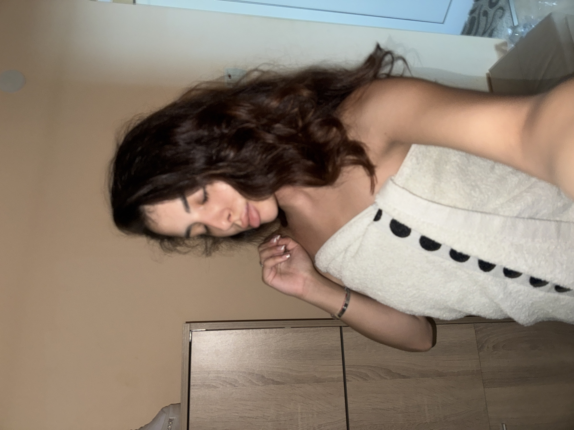 A young woman with dark, wavy hair is pictured from the chest up, wrapped in a white towel with black polka dots. She is looking down with her eyes closed and her hand resting on her chest. The background is a plain, beige wall, with a wooden cabinet to her left and a doorway to her right. There is no indication of the city of Plovdiv, Bulgaria.