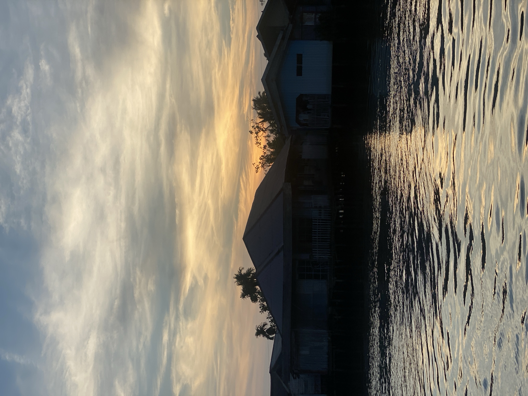 The image depicts a scene in Banjarmasin, Indonesia, at dusk, likely along one of the city's many rivers or canals. The foreground is dominated by rippling water reflecting the warm hues of the setting sun. In the mid-ground, a row of stilt houses lines the water's edge, silhouetted against the sky. The architecture suggests traditional Indonesian vernacular housing, with prominent pitched roofs and raised foundations. Palm trees are visible amongst the houses, characteristic of the tropical climate. The sky above is a canvas of wispy clouds, transitioning from a clear blue at the top to soft oranges and yellows near the horizon, indicating the sun is setting. There are no people visible in the image.