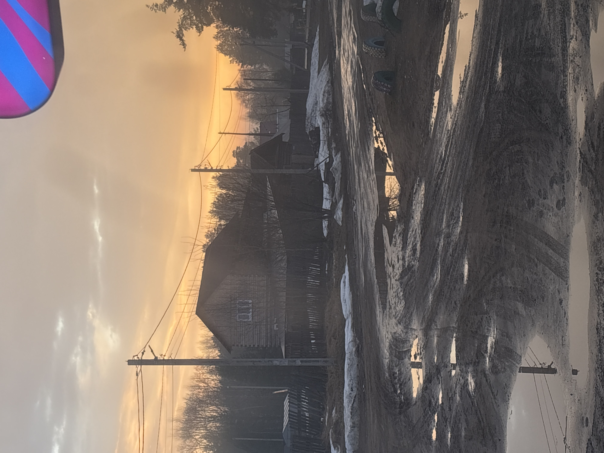 The image shows a rural scene, possibly in or near Rusalevka, Russia, given the prompt context. A weathered wooden building, likely a house, stands in the mid-ground. Power lines stretch across the sky, connecting to poles alongside the building. The ground is a mix of muddy and snowy patches, suggesting a transition season. A road or path is visible with tire tracks and puddles reflecting the sky. In the top left corner, there is a portion of a colorful, striped object, possibly a hot air balloon. The overall lighting indicates either sunrise or sunset, casting a warm glow in the sky and creating long shadows. The image has a slightly desaturated quality.