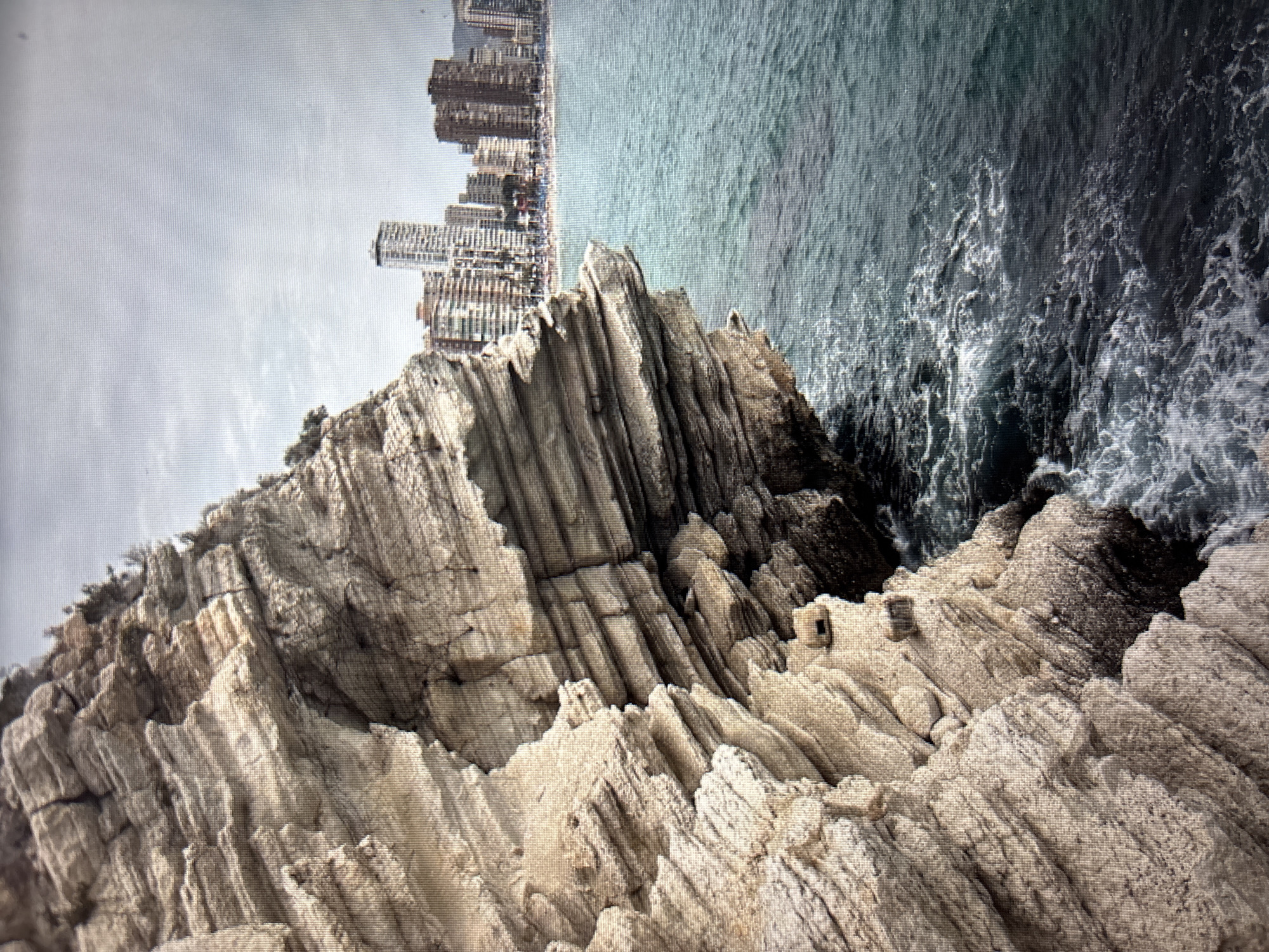 The image depicts a rocky cliff face on the left side, with layers of weathered stone. To the right of the cliff is the ocean, with choppy, dark blue water and white foam. In the distance, a city skyline is visible, characterized by numerous tall buildings, including some that appear to be skyscrapers. The sky is overcast and a pale gray. The scene is outdoors and appears to be a coastal area with an urban backdrop. This location is not in Sofia, Bulgaria.
