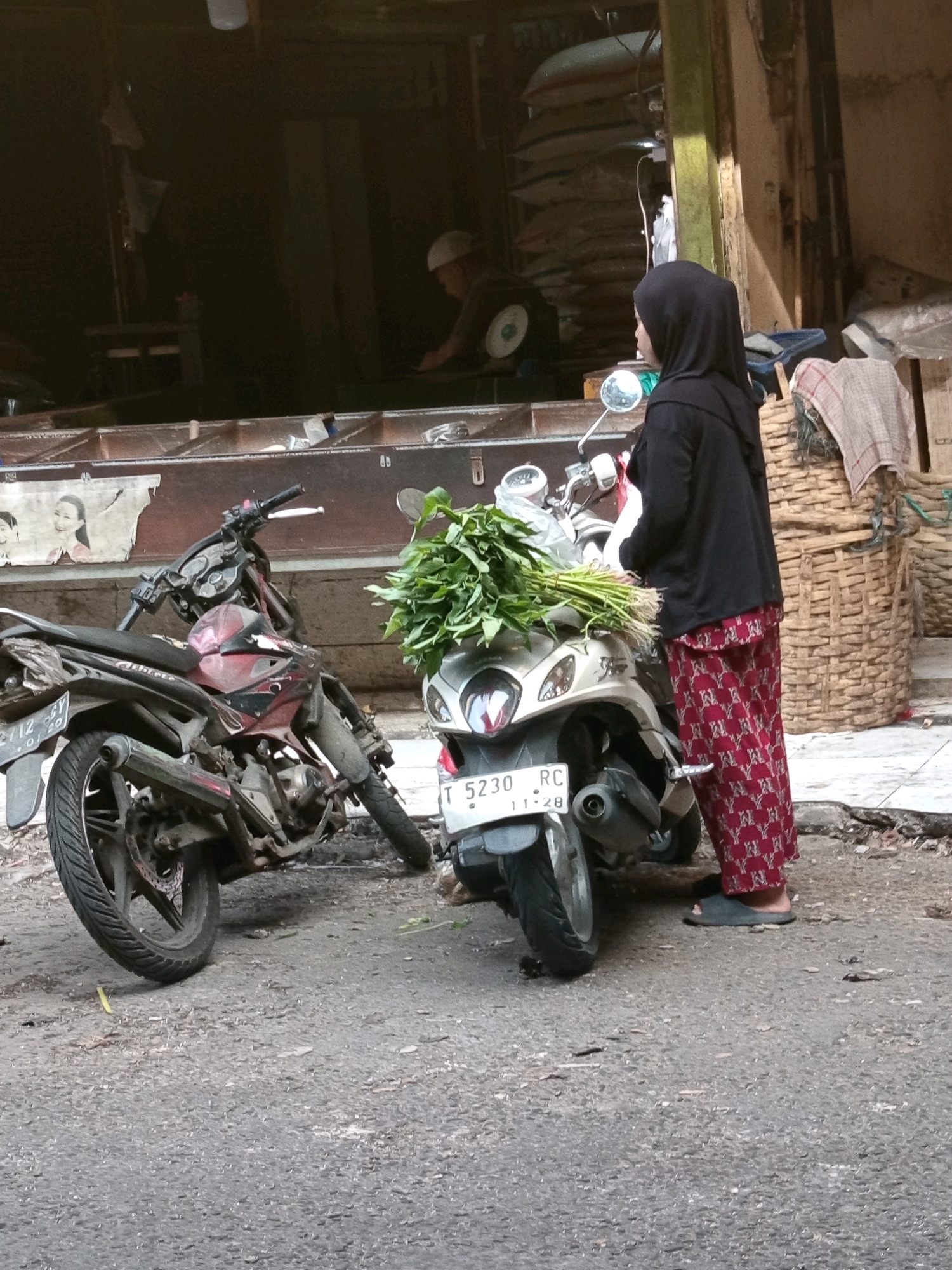 A woman wearing a black hijab and red patterned pants stands next to a white scooter. The scooter has a large bunch of green leafy vegetables tied to its front. To the left of the white scooter is a red and black motorcycle. The scene appears to be a street in Karawang, Indonesia, with a building in the background that may be a market or shop. The ground is paved with asphalt.