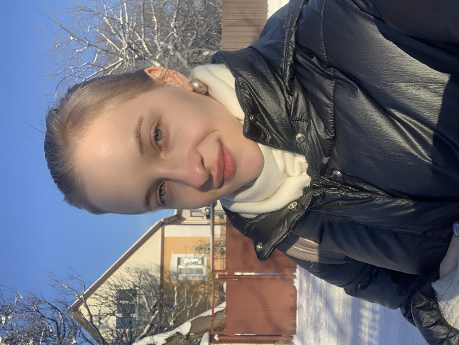 An outdoor scene in Bohuslav, Ukraine, depicts a young female in a close-up, selfie-style shot during winter. The subject, positioned centrally, is a fair-skinned female with light-colored eyes and light brown hair pulled back. She exhibits a subtle smile and looks directly towards the camera. She wears a black puffer jacket, an off-white scarf, and a large, light-colored stud earring on her right ear.
The environment is a clear, sunny winter day, evidenced by a bright blue sky and snow covering the ground, bare tree branches, and fence tops. In the background, to the left of the subject, stands a two-story residential house with light yellow siding and a light-colored roof, featuring a visible window on its first floor. A reddish-brown metal gate, likely part of a fence, is situated in front of the house. To the far right, a section of a light brown wooden fence is visible. Bare trees, laden with snow, are present on both sides of the background. Bright sunlight casts distinct shadows across the scene and illuminates the subject's face.