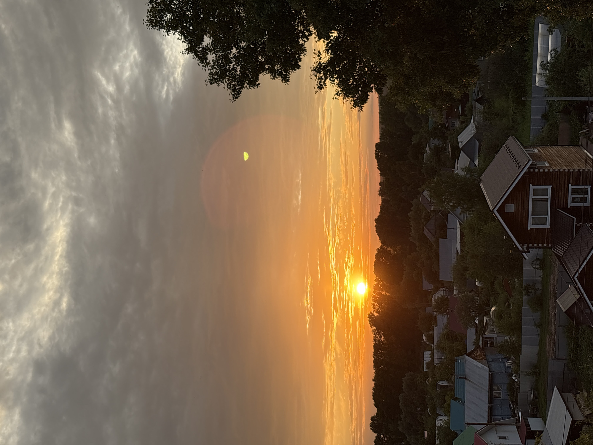 The image depicts a sunset scene over a residential area in Chaikovsky, Russia. The sun, a bright, glowing orb, is low on the horizon, casting a warm orange and yellow light across the sky. Wispy clouds are scattered throughout, some catching the sunlight and appearing to glow. The sky transitions from a bright, fiery orange near the sun to a softer, hazy grey towards the left of the frame.
In the foreground and mid-ground, a cluster of houses and buildings is visible, nestled amongst lush green trees. The architecture suggests a rural or suburban setting, with many houses appearing to be wooden structures with pitched roofs. Some of the buildings have visible windows and chimneys. The trees are dense and leafy, indicating it is likely summer or early autumn.
The overall mood of the image is peaceful and serene, characteristic of a quiet evening at sunset. The vibrant colors of the sky and the silhouette of the trees and houses create a picturesque and tranquil atmosphere. The specific location cues like the style of houses and the vegetation point towards a Russian setting. The time of day is clearly sunset, given the position and color of the sun. There is no visible text in the image.