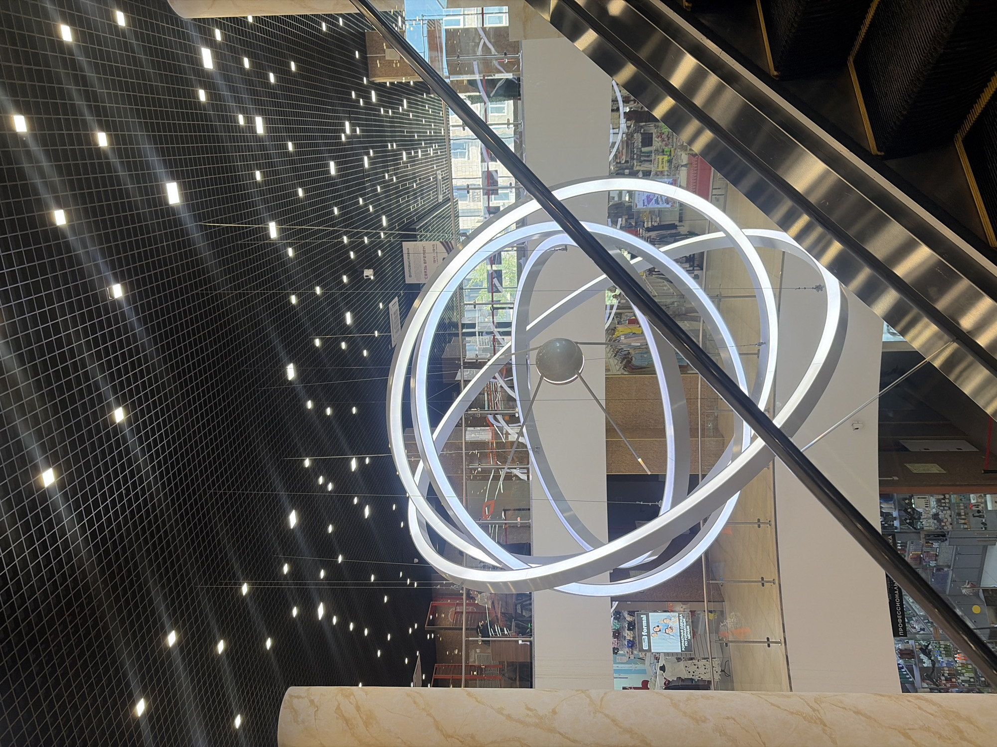The image shows the interior of a shopping mall in Saint Petersburg, Russia. The view is upwards, showcasing a modern lighting fixture comprised of concentric illuminated rings, suspended from the ceiling. Adjacent to the lighting, an escalator is visible, with its metallic structure and moving steps. The mall features a grid-patterned wall or ceiling with embedded lights, creating a dispersed glow effect. A pillar with a mottled, marbled pattern is visible in the foreground. The mall architecture incorporates glass panels and reflective surfaces, allowing for multiple perspectives and views of internal shops and building structure.